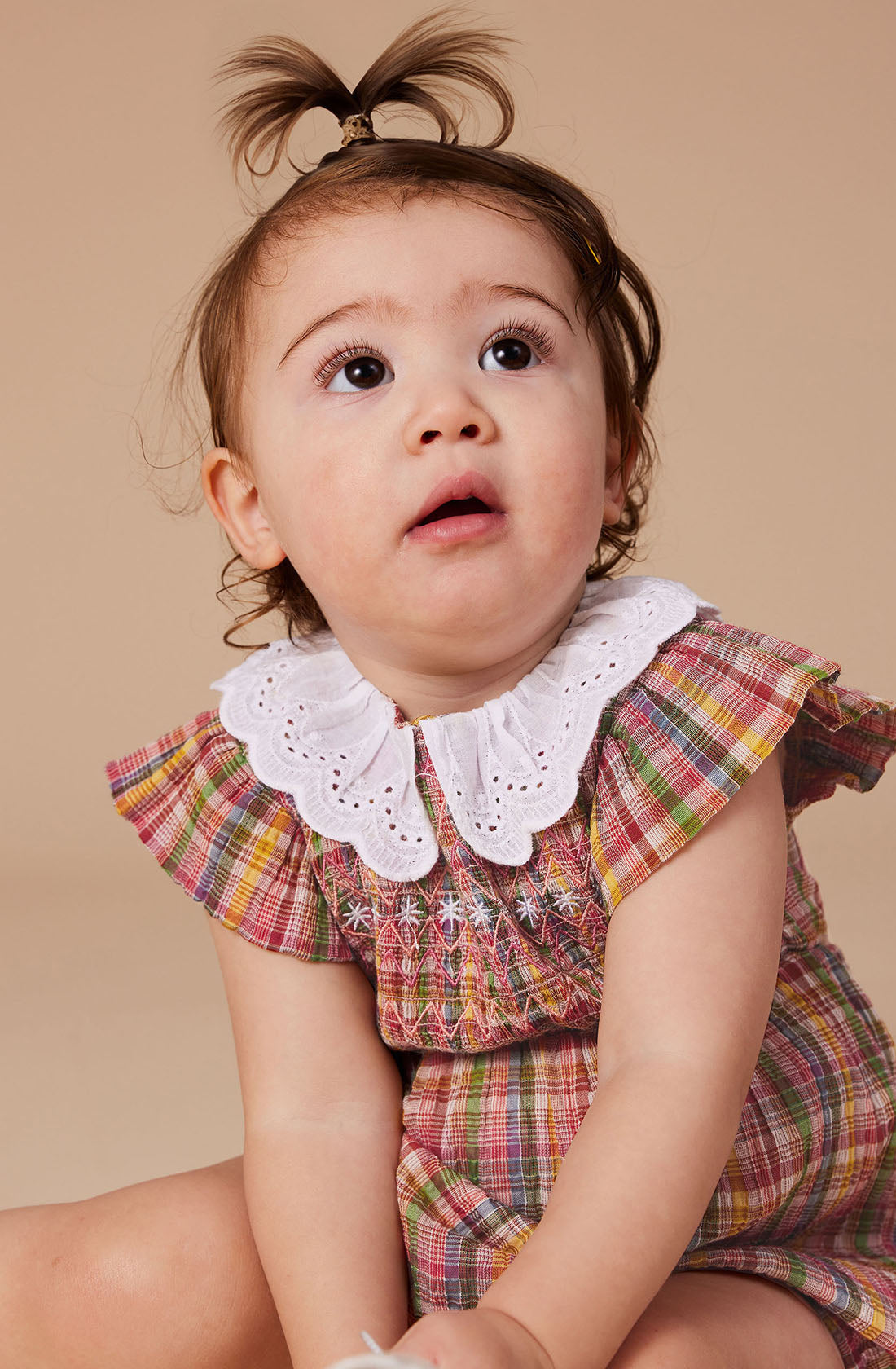 A young child sits against a plain beige background, looking slightly upward. The child has a small ponytail on top of their head and is wearing the colorful Flo Smocked Romper by GOLDIE + ACE, featuring a smocked bodice and a charming white eyelet lace collar. The romper also has elastic balloon sleeves that add to the adorable look.