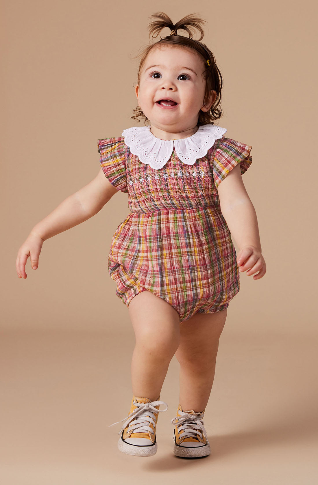 A light brown-haired toddler stands wearing the vibrant Flo Smocked Romper by GOLDIE + ACE, which boasts a colorful plaid design and a charming white lace collar, perfectly complemented with matching yellow and white shoes. The romper features delightful elastic balloon sleeves. The child's small ponytail is adorned with a yellow clip as they smile while looking slightly upward against a plain beige background.