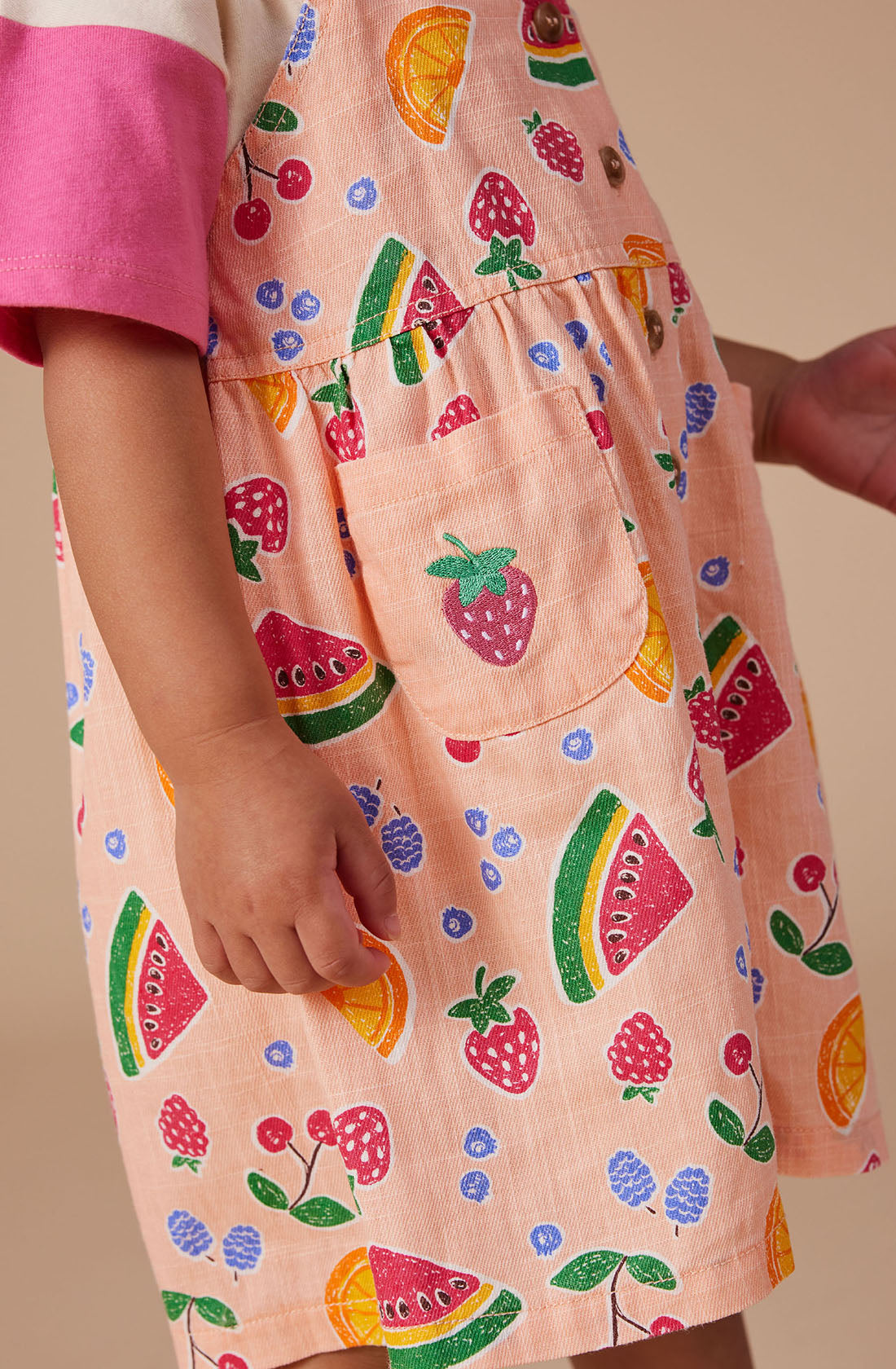 A child wearing GOLDIE + ACE's Sarah Linen Shortalls, crafted from a colorful linen/cotton blend and adorned with a playful hand-drawn fruit print featuring watermelons, cherries, strawberries, lemons, and blueberries. The shortalls have adjustable button straps and a small pocket with a strawberry design. The child is pictured from the shoulders down with their arm slightly extended.