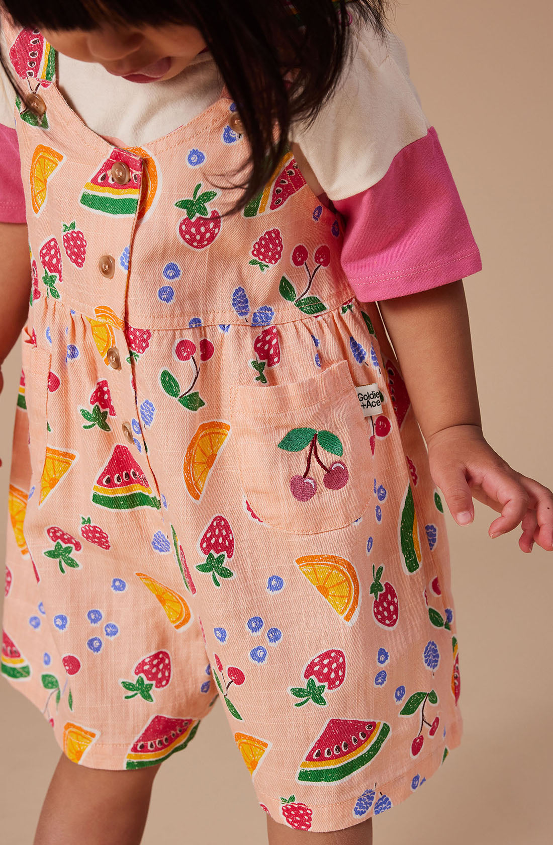 A young child wearing the Sara Linen Shortalls by GOLDIE + ACE, adorned with hand-drawn prints of strawberries, blueberries, watermelons, and oranges in vibrant colors. The linen and cotton blend shortalls feature adjustable button straps. The child is also sporting a white shirt with pink sleeves underneath and is gazing slightly downward.