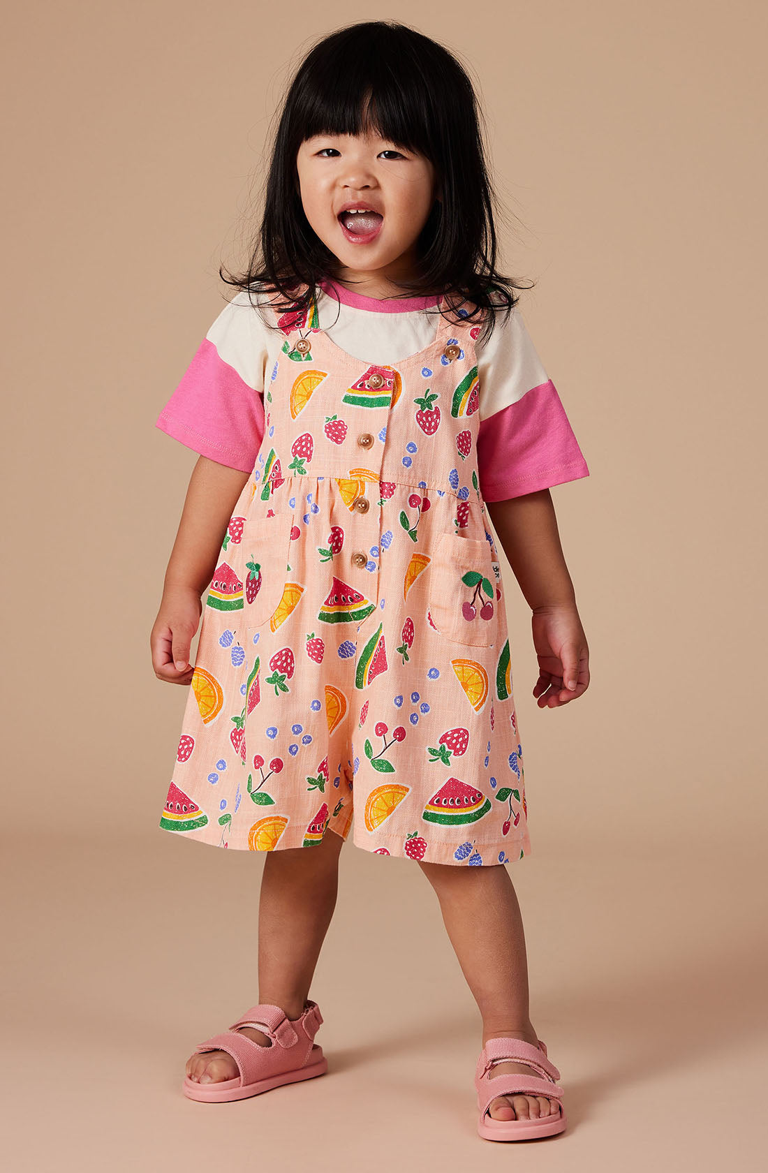 A young child with long, dark hair stands against a beige background, wearing the Sara Linen Shortalls by GOLDIE + ACE, which feature colorful fruit patterns and adjustable button straps, along with a pink and white T-shirt underneath, and pink sandals. The child's open mouth suggests they may be speaking or singing.