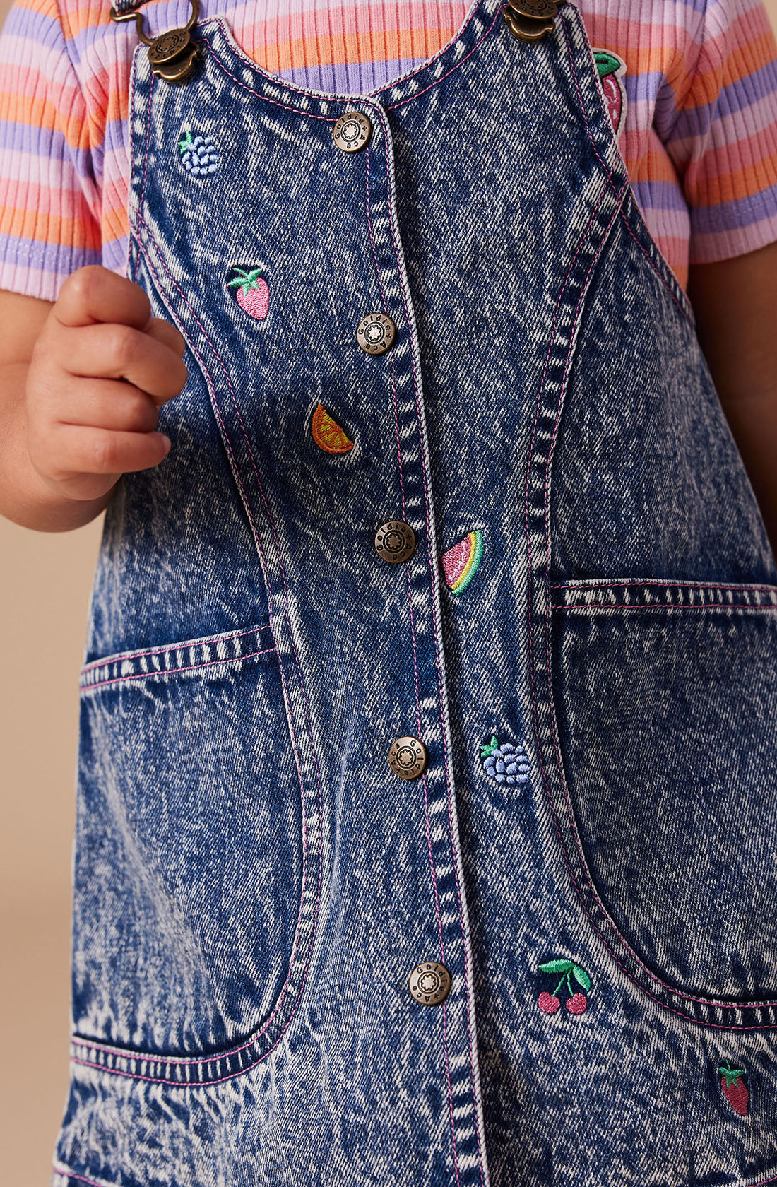 A close-up shows a child wearing the Penelope Fruity Denim Pinafore Dress by GOLDIE + ACE. The dark denim dress is adorned with hand-drawn, embroidered fruit patches featuring strawberries, cherries, and a slice of watermelon. It features adjustable brass straps, a front button closure, and two front pockets. The dress is layered over a striped shirt.