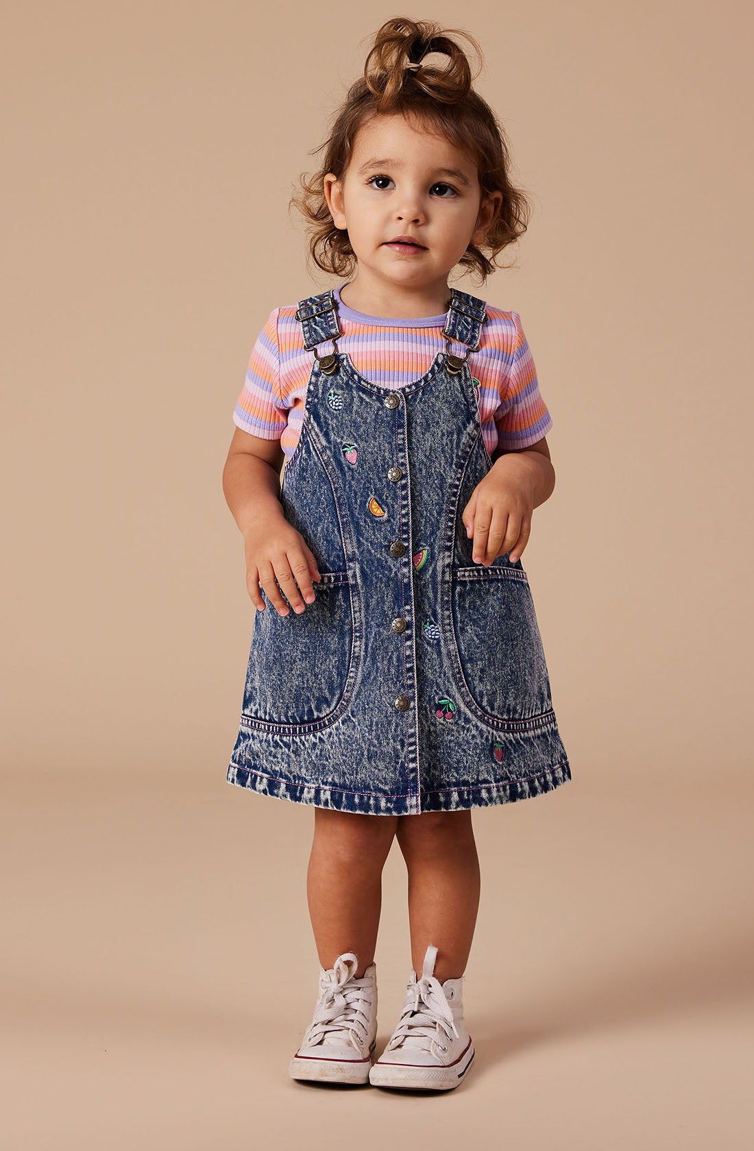 A young girl with medium-length curly hair stands against a plain background. She wears a striped short-sleeved shirt under the Penelope Fruity Denim Pinafore Dress from GOLDIE + ACE, which features colorful patches and adjustable brass straps. She completes her outfit with white sneakers and looks directly at the camera with a neutral expression.