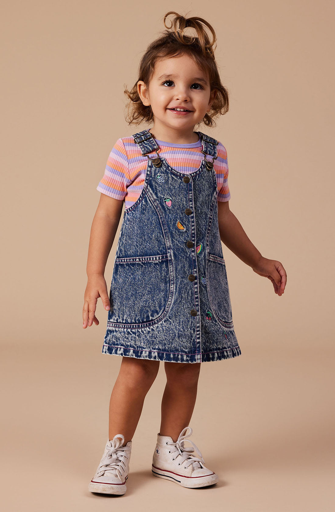 A smiling toddler stands on a beige background, wearing a striped short-sleeve shirt under the Penelope Fruity Denim Pinafore Dress by GOLDIE + ACE. The dress features adjustable brass straps and colorful heart patches, perfectly complemented by her white sneakers. Her hair is partially pulled up into a small ponytail on top of her head.