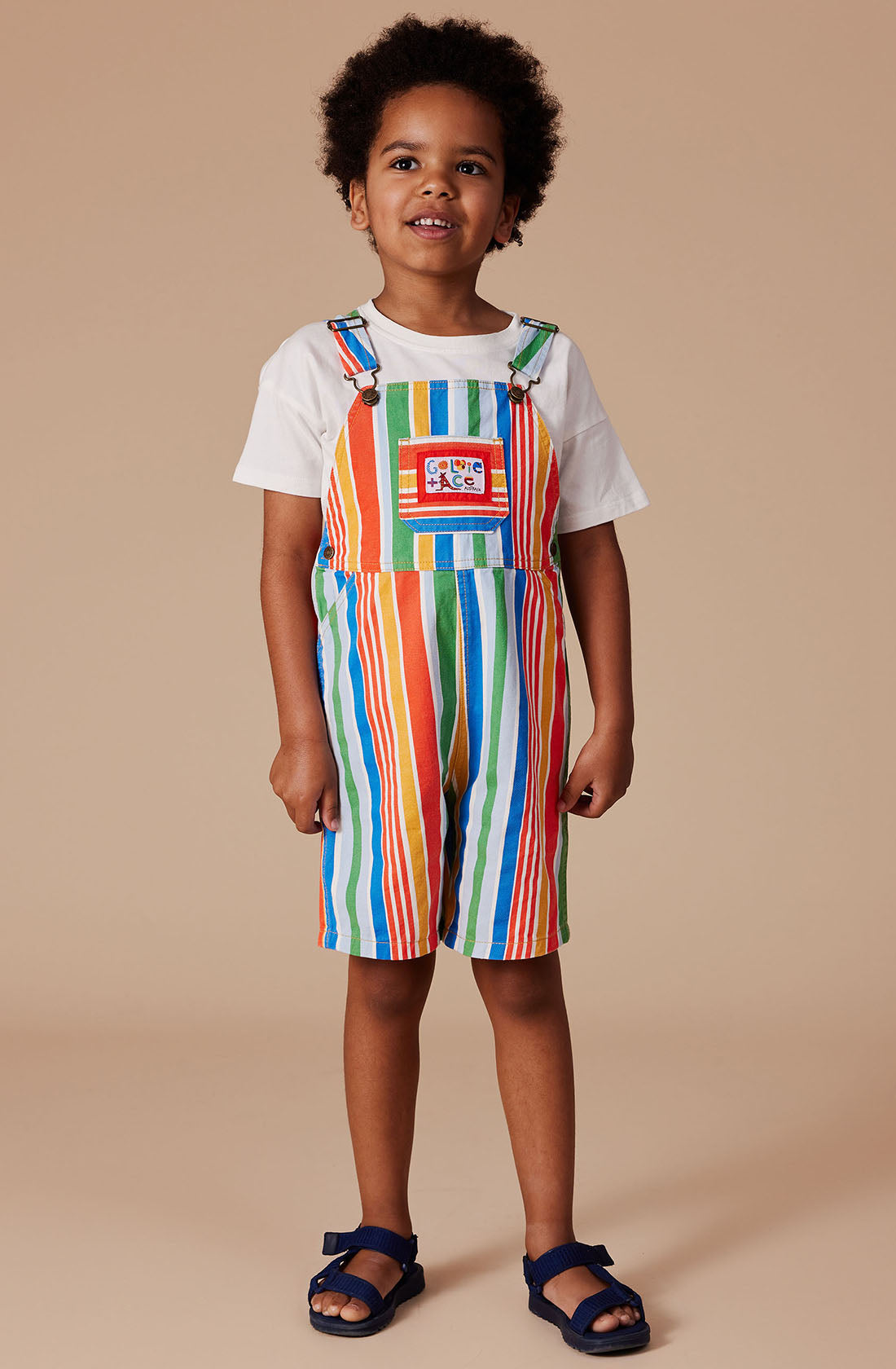 A child smiles, dressed in the vibrant Burton Striped Denim Overalls by GOLDIE + ACE, paired with a white T-shirt underneath. The denim overalls feature vertical stripes in green, orange, blue, and white, complete with adjustable straps and a small patch on the chest. The child stands against a plain beige background, wearing navy blue sandals.