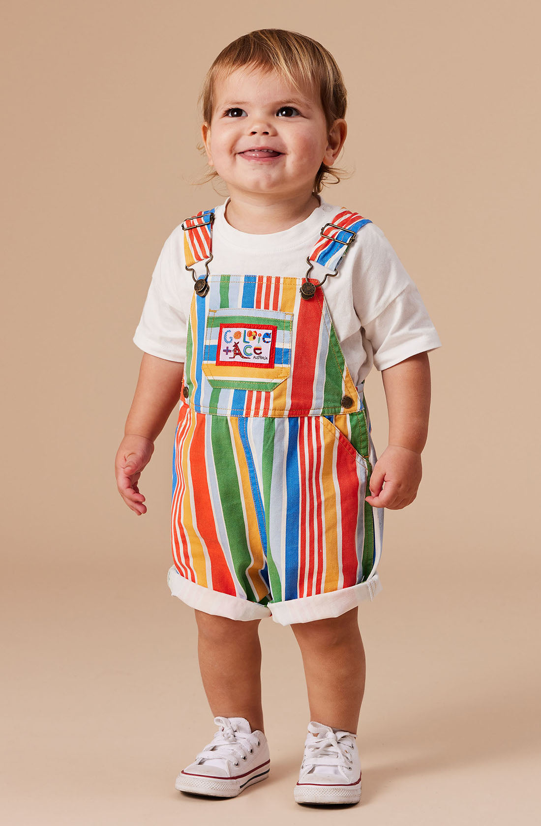 A toddler with short hair smiles while wearing a white t-shirt underneath Burton Striped Denim Overalls by GOLDIE + ACE. The red, green, yellow, and blue striped overalls feature a playful pocket design and leg snaps for easy changes. The child is also wearing white sneakers.