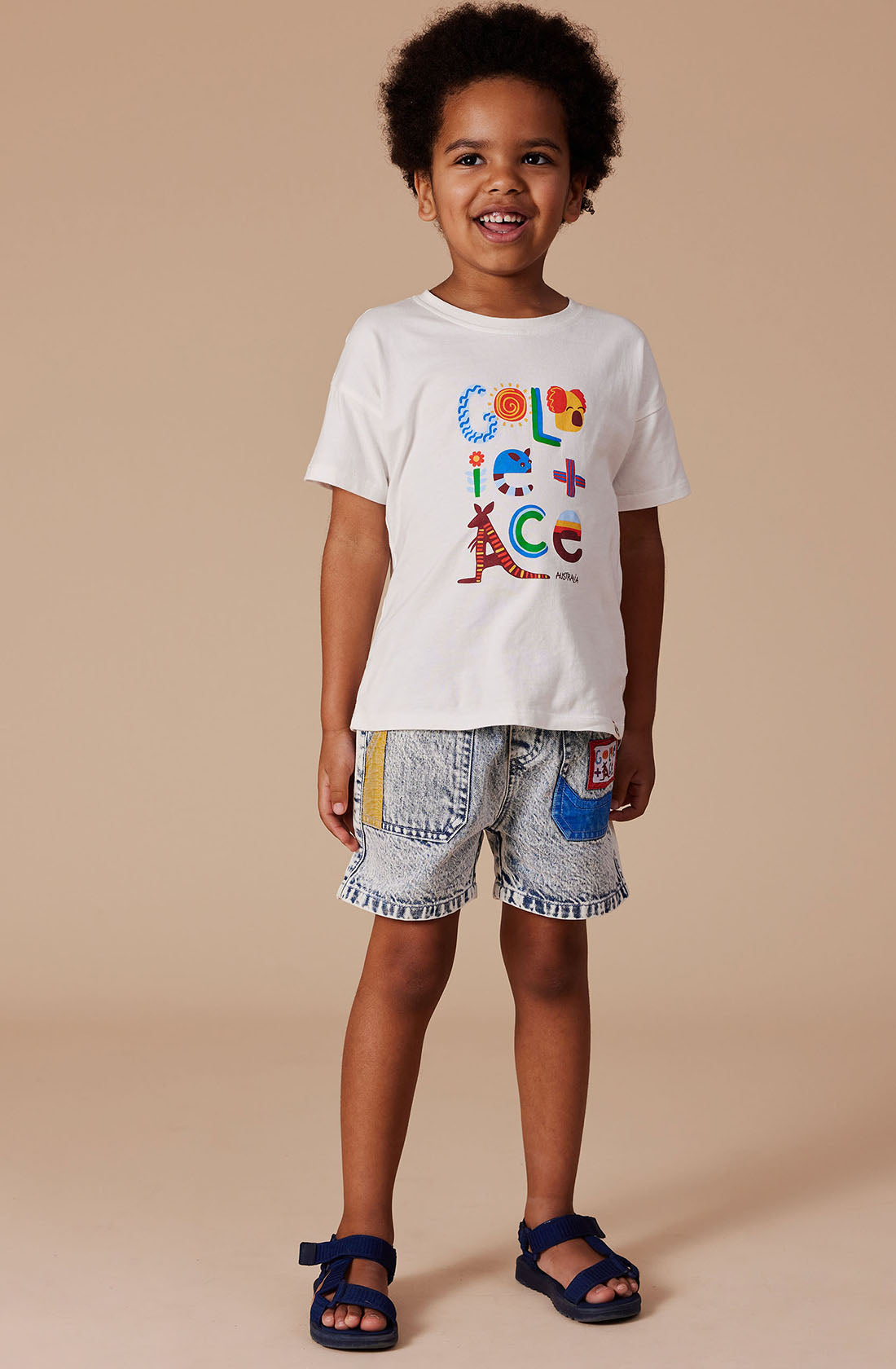 A young child with curly hair stands smiling against a beige background. They are wearing a white t-shirt with colorful text that reads "Color is ace" and GOLDIE + ACE's light denim Noah Colour Pop Denim Shorts, which feature patches and an elastic waistband. Blue sandals complete their outfit.