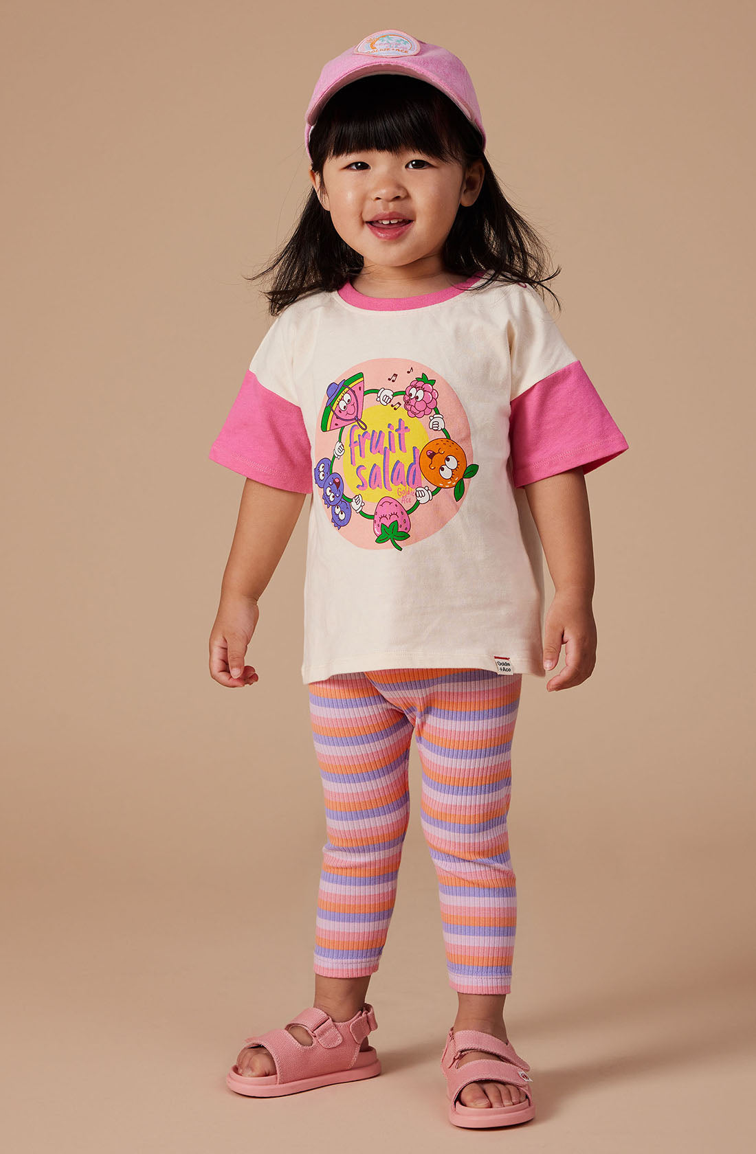 A young girl with dark hair stands smiling against a plain beige background. She is wearing a light pink baseball cap, GOLDIE + ACE's Fruit Salad Friends T-shirt featuring pink sleeves and a hand-drawn "Fruit Salad" graphic, super soft stretch cotton jersey pink and blue striped leggings, and pink sandals.