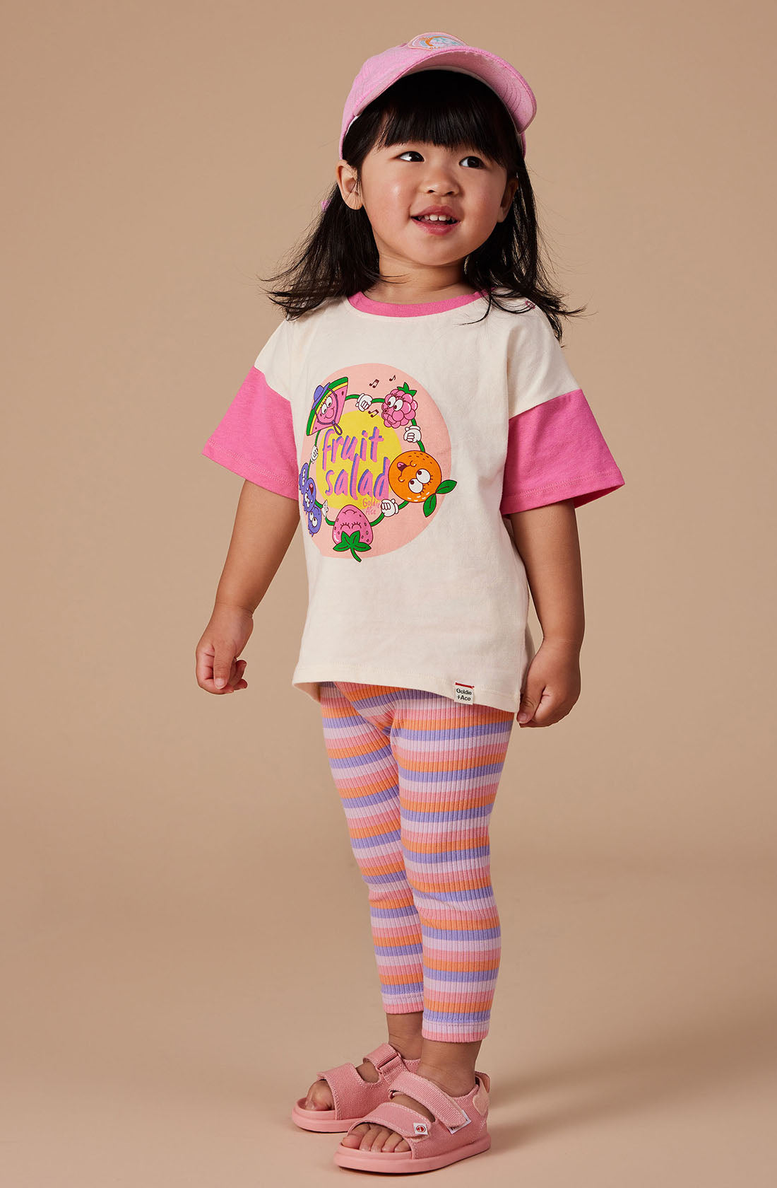 A young child with shoulder-length dark hair is wearing a pink baseball cap, a white and pink t-shirt from GOLDIE + ACE's "Fruit Salad Friends" collection, striped leggings in pink and orange, and pink sandals. The background is plain beige. The super-soft stretch cotton jersey t-shirt perfectly complements the child's cheerful smile.