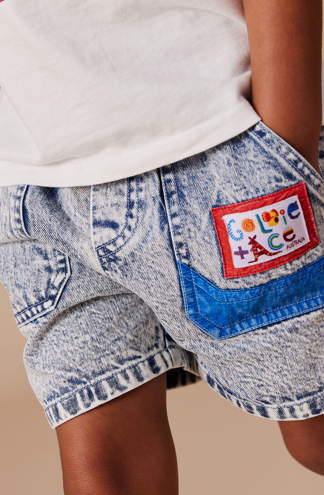 A close-up shot of a person wearing the Noah Colour Pop Denim Shorts from GOLDIE + ACE. The light denim shorts feature a red-bordered patch on the back pocket, adorned with word graphics such as "Love" and various small icons like a smiley face, Eiffel Tower, and other playful designs. The shorts also come equipped with a functional drawcord for added convenience.