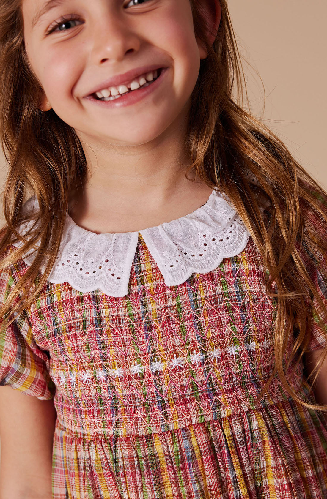 A young girl with long hair smiles joyfully, dressed in the FLO Smocked Dress by GOLDIE + ACE. The dress features intricate multicolored embroidery on a plaid fabric and a white lace collar, accentuated by a smocked bodice. The neutral background ensures that her cheerful expression and beautifully detailed outfit remain the focal points.