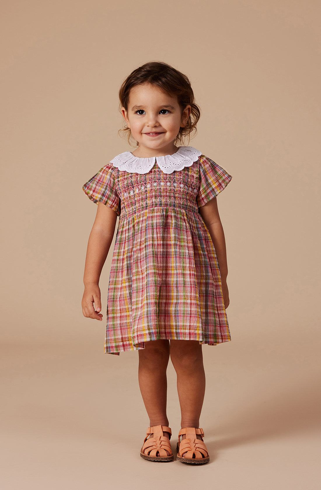 A young child stands against a neutral background, smiling and looking at the camera. She is wearing the vibrant Flo Smocked Dress from GOLDIE + ACE, featuring short sleeves, a white Peter Pan collar, and a smocked bodice. Her outfit is paired with tan sandals, and her curly hair is tied back.