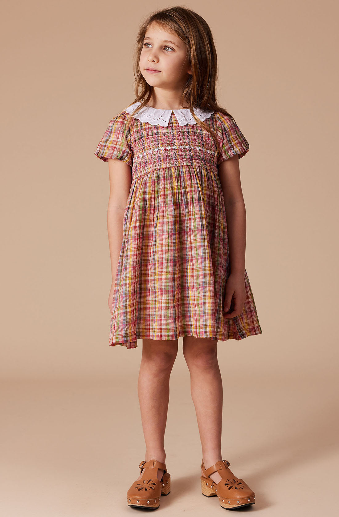 A young girl with long brown hair stands against a plain background, wearing the Flo Smocked Dress from GOLDIE + ACE. The colorful plaid dress features a white lace collar, puffed sleeves, and a smocked bodice. The Maroon Rainbow check pattern of the crinkle cotton fabric adds a charming touch. She has brown shoes with cut-out patterns and gazes slightly to her right.