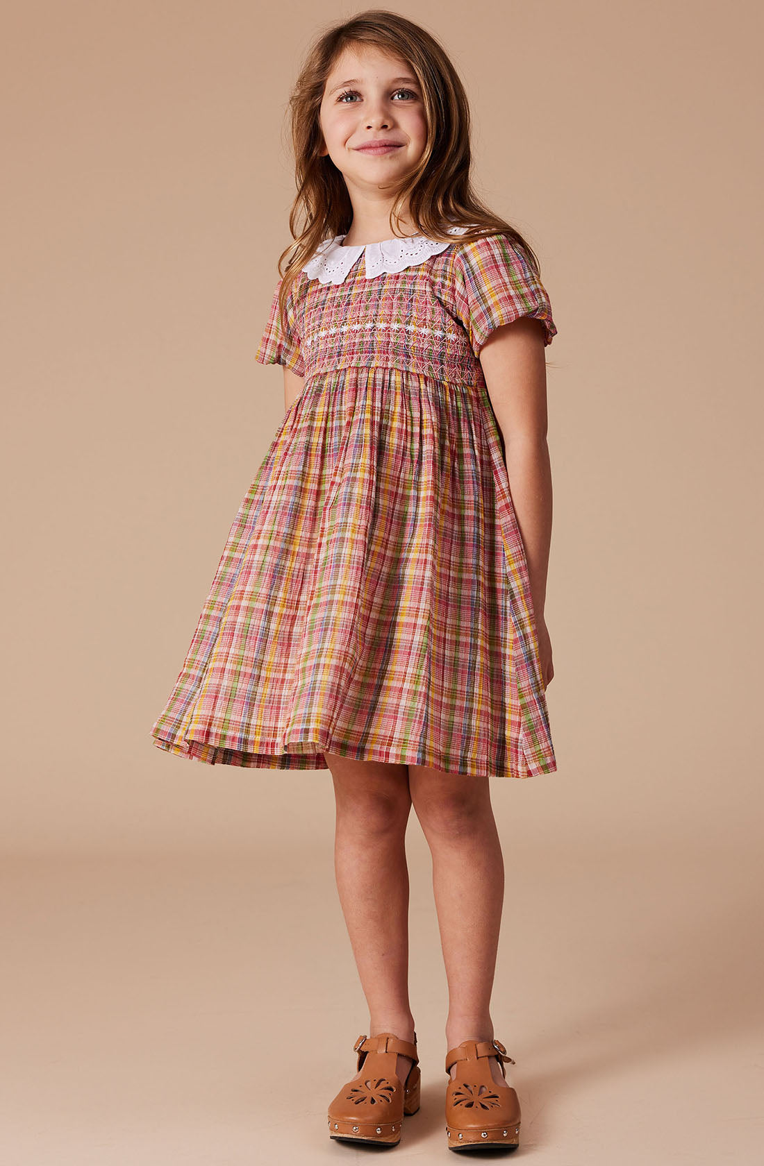 A young girl with long brown hair is standing and smiling, wearing the Flo Smocked Dress by GOLDIE + ACE. The dress features a maroon rainbow check plaid pattern with a white Peter Pan collar, and she has paired it with brown shoes. The background is plain and light-colored.