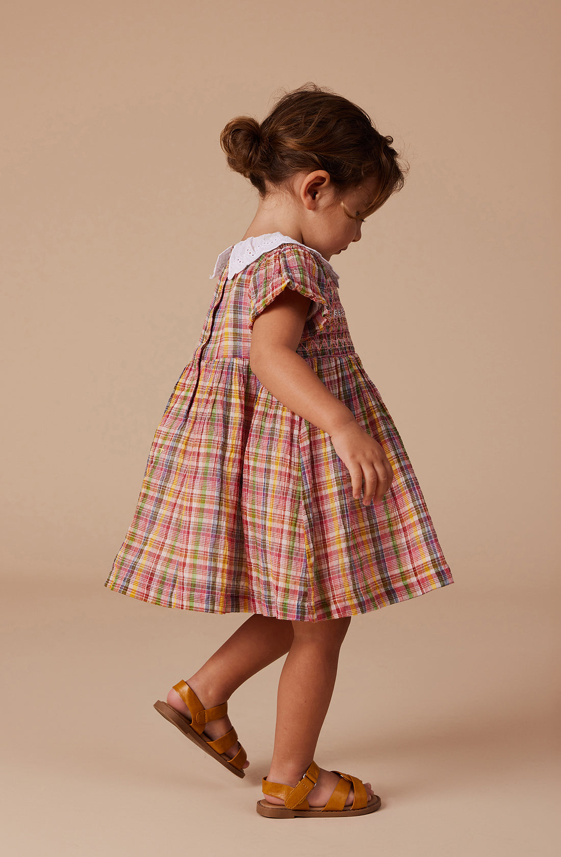 A young child wearing the "Flo Smocked Dress" by GOLDIE + ACE, featuring a colorful plaid pattern and a white lace collar, stands against a neutral beige background. She pairs the dress with brown sandals, and her hair is tied back in a low bun. The short sleeves, gathered skirt, and smocked bodice of the dress add charm to the scene.