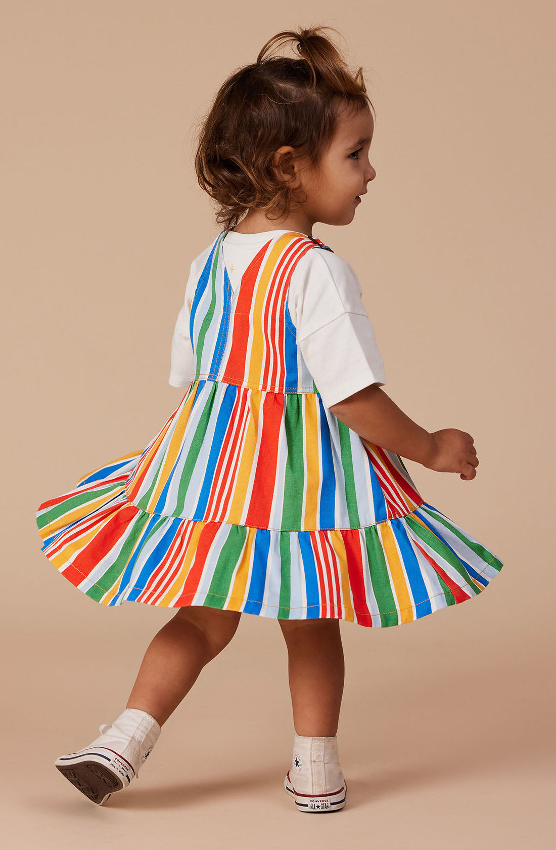 A toddler with light brown hair stands facing sideways. The child is wearing a colorful Tully Tiered Pinafore Dress from GOLDIE + ACE, featuring Red/Green/Yellow/Blue stripes, adjustable straps, and brass snap buttons. They are also sporting white shoes with a blue logo. The background is a plain, light beige.