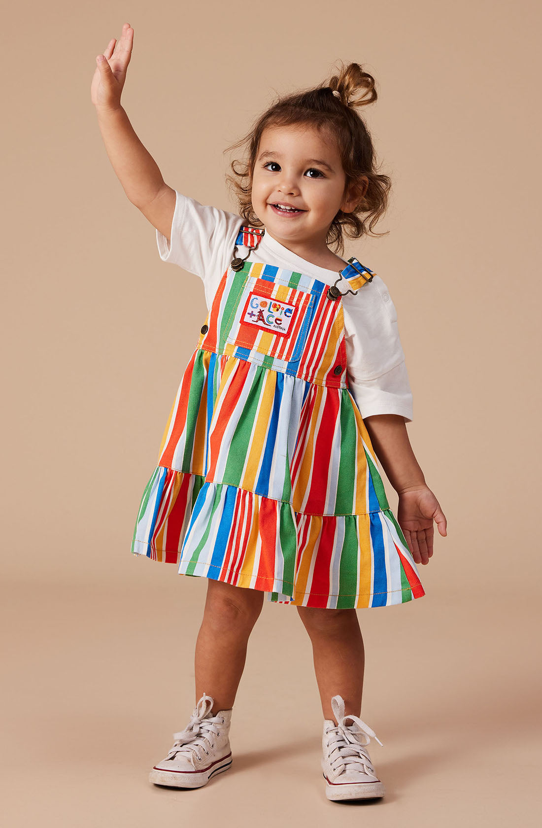 A young child with brown hair tied in a small ponytail, wearing the vibrant Tully Tiered Pinafore Dress by GOLDIE + ACE and white sneakers, stands against a beige background. The child smiles and waves with one hand raised.