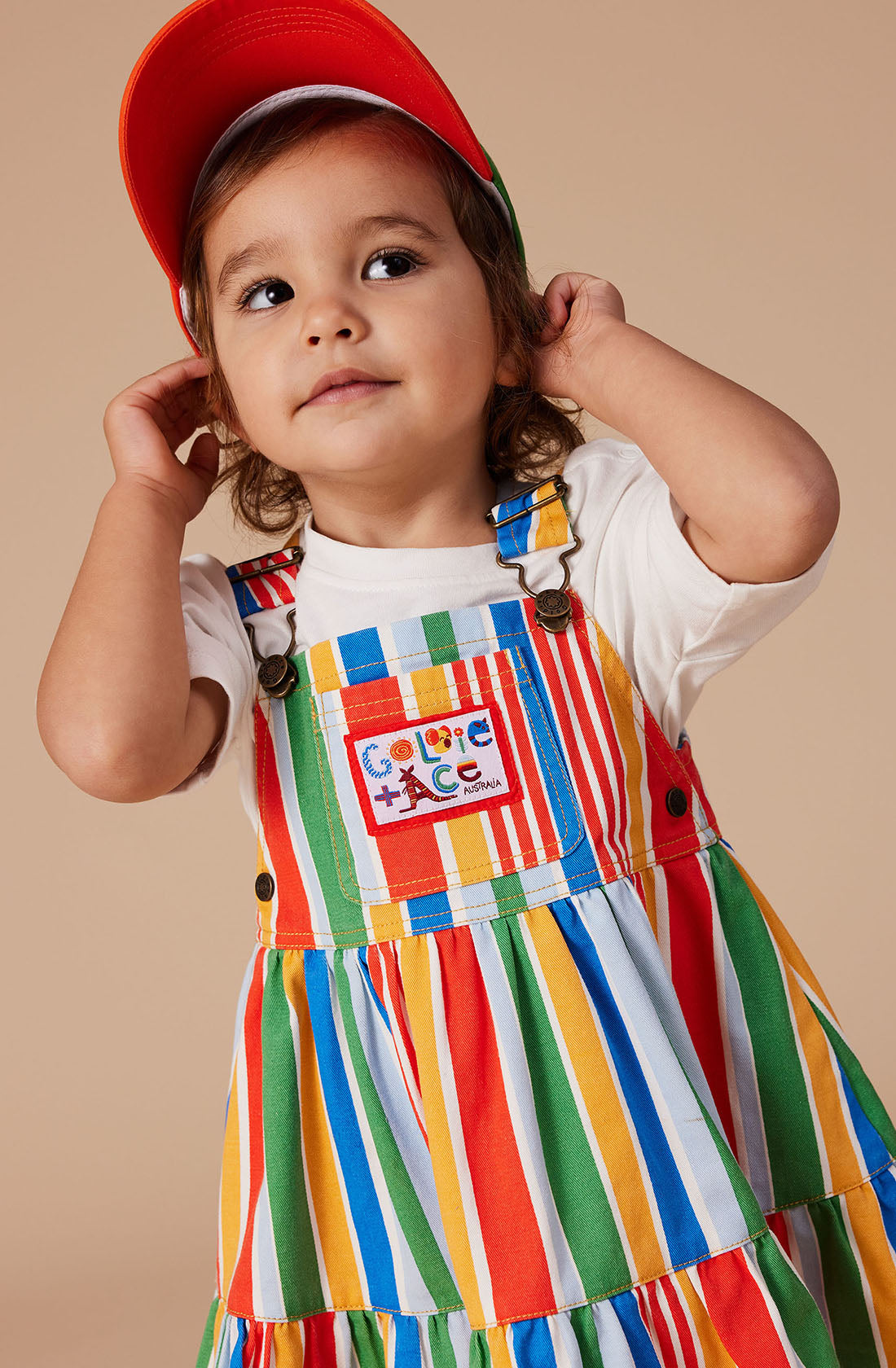 A young child with long hair wears a red cap and a white t-shirt underneath the colorful Tully Tiered Pinafore Dress by GOLDIE + ACE. The dress features vertical stripes in red, green, yellow, and blue, brass snap buttons on the adjustable straps, and a patch on the front pocket. The child looks off to the side with hands near their ears.