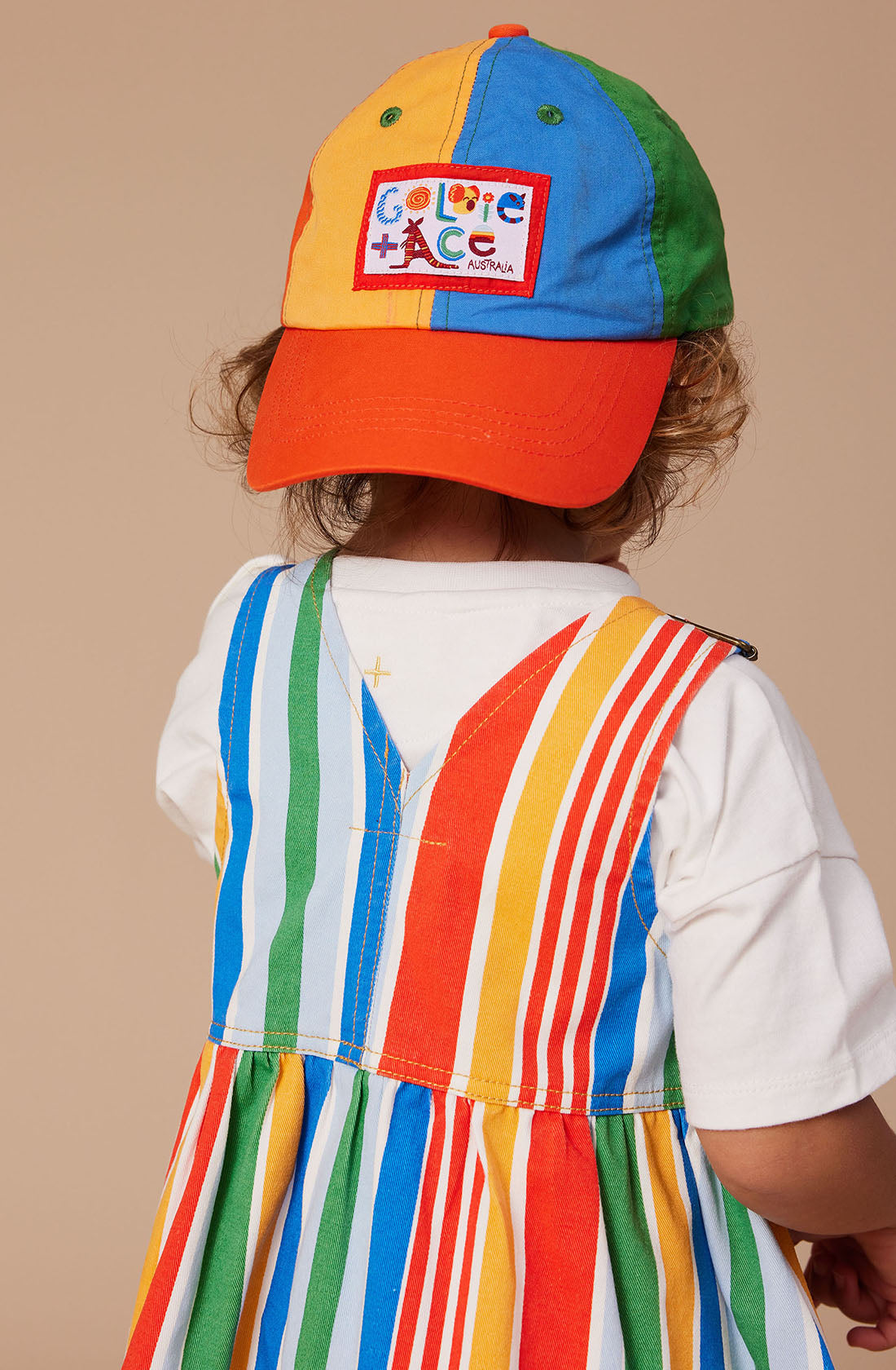 A toddler is seen from behind wearing a colorful, striped jumper and the Tate Land Down Under Denim Cap by GOLDIE + ACE, which comes with an adjustable strap. The dress and cap boast vibrant vertical stripes in blue, green, yellow, orange, and red. The cap also features a label with text. The background is a neutral beige color.