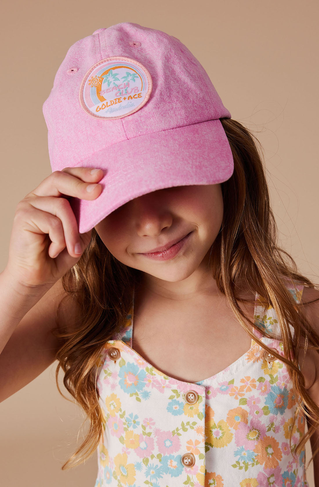 A young girl is captured wearing a Tate Beach Club Denim Cap by GOLDIE + ACE, the cap's light pink shade and embroidered logo slightly obscuring her face. It showcases a washed denim finish and an adjustable strap for a perfect fit. Paired with her floral-patterned, sleeveless top adorned with front buttons, her long wavy hair cascades over her shoulders as she smiles slightly and tilts the cap.