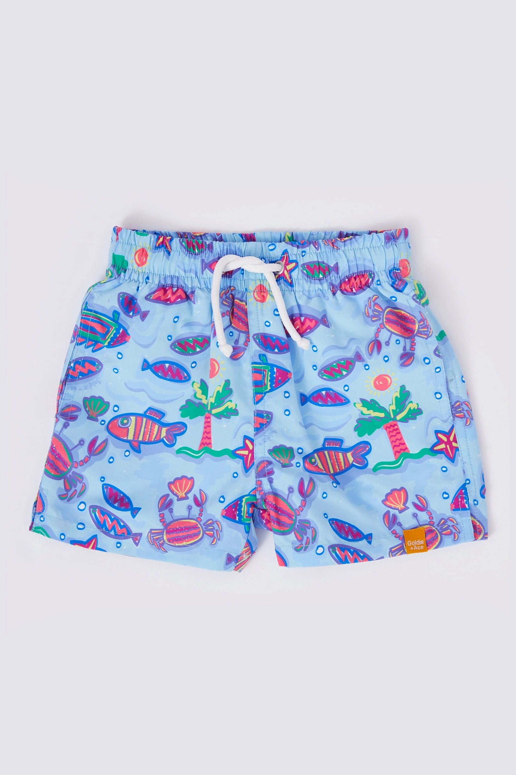 GOLDIE + ACE's Daydream Island Board Shorts are light blue children's swim trunks made from soft Microfibre Twill, showcasing vibrant, colorful aquatic-themed prints of fish, coral, and plants. These shorts feature a functional drawstring on the elastic waistband and a small orange tag on the hem of the right leg.