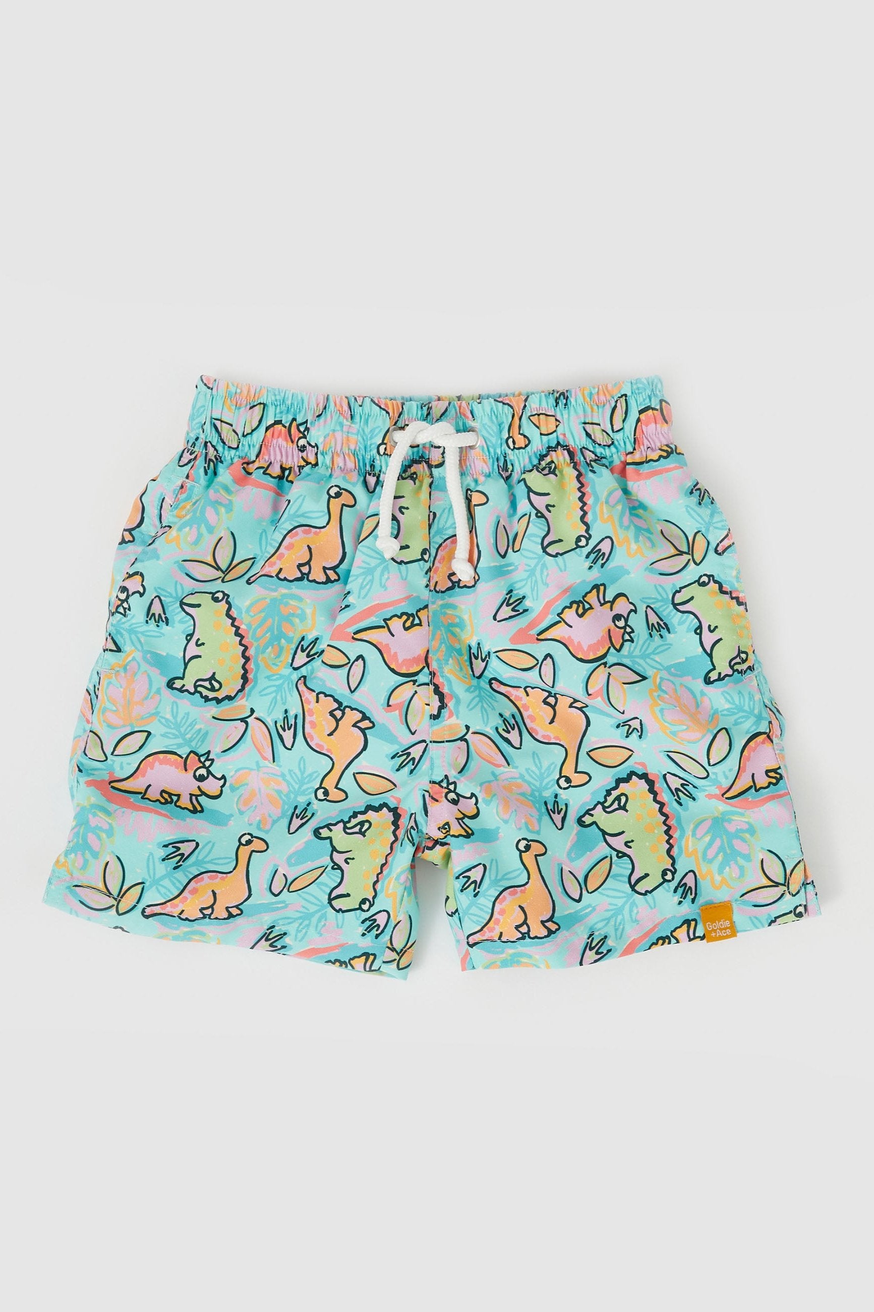 The GOLDIE + ACE Dino Jungle Board Shorts are a pair of colorful swim trunks adorned with various cartoon dinosaur illustrations on an aqua green multi microfiber twill. The design features green, pink, and yellow dinosaurs set against a light blue background, along with leaves and other tropical elements. They are true to size with a white drawstring and an elastic waistband.