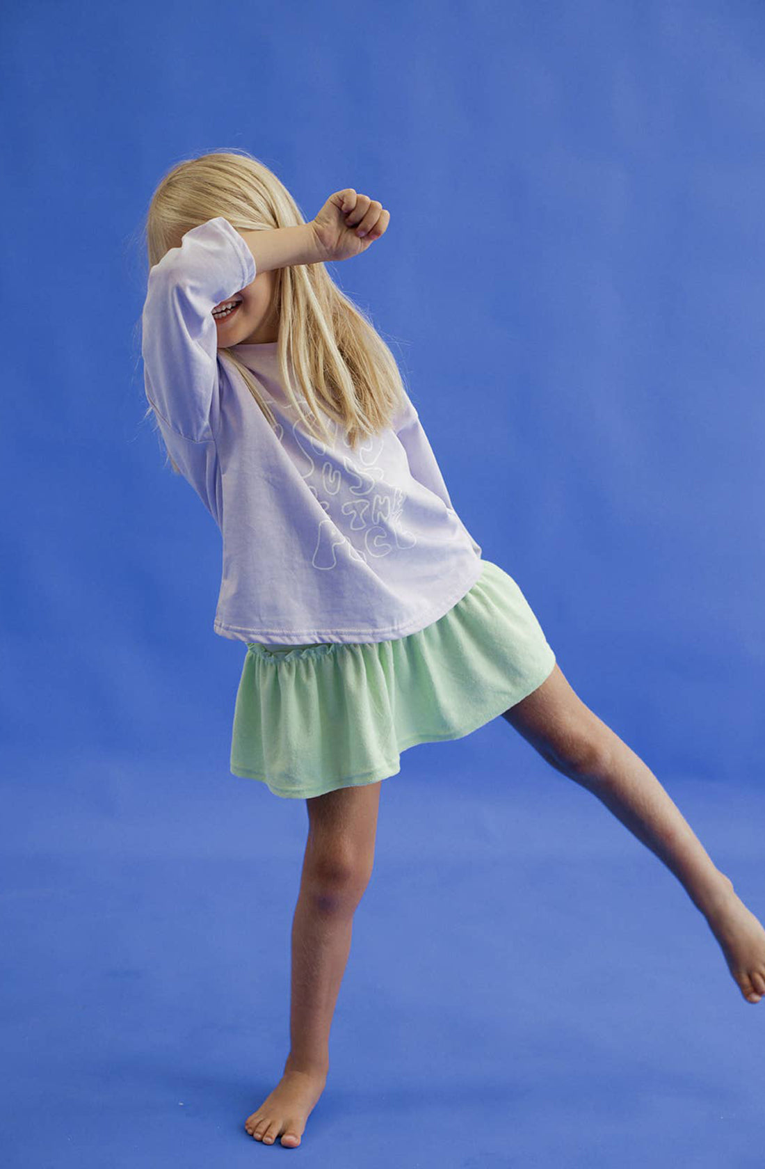 A young girl wearing the spin me round terry towelling dress in green and a purple long sleeved top. She is covering her eyes with her hand.