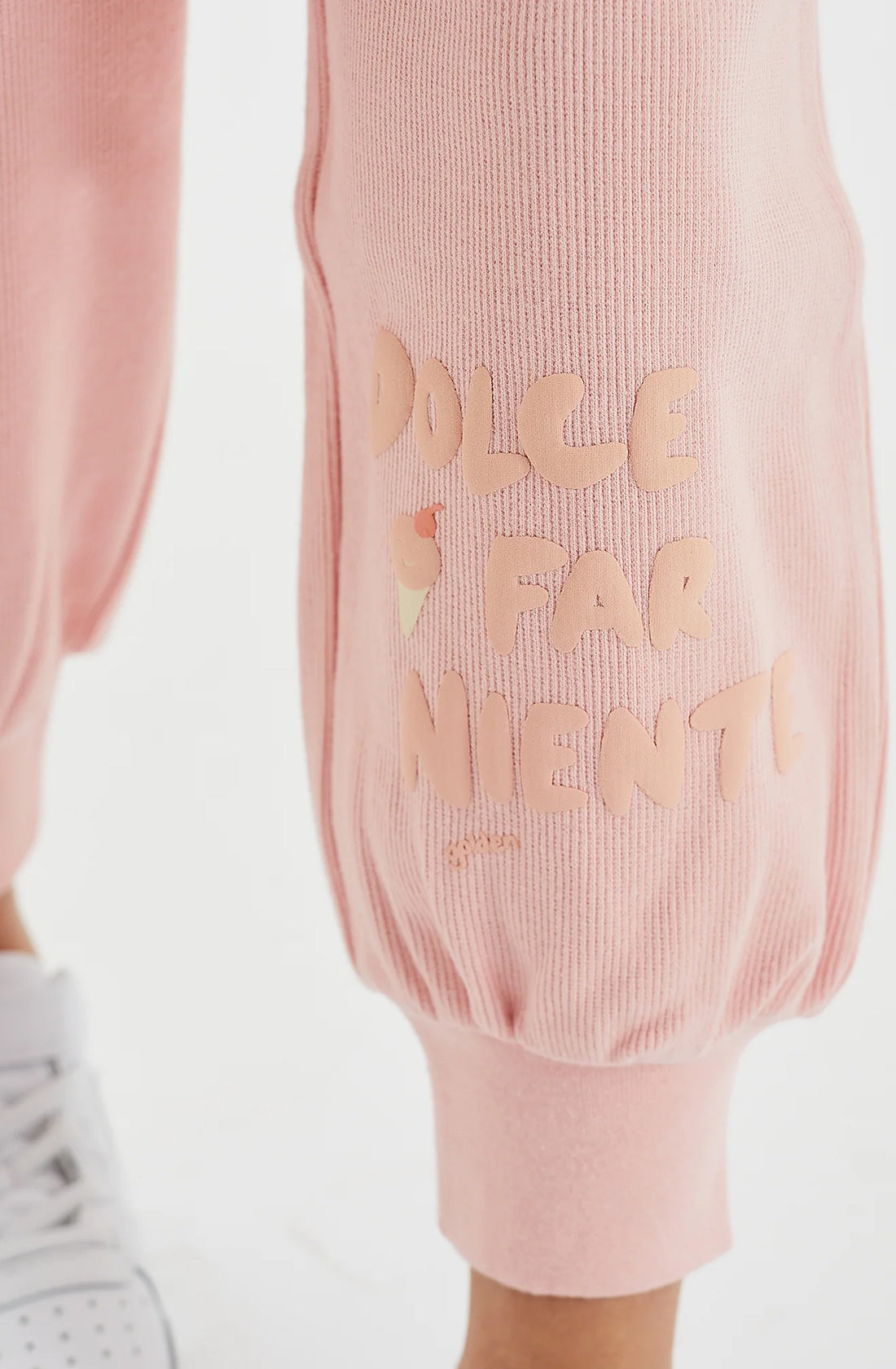 Close-up of a person wearing light pink Dolce Lounge Pant Pink Lemonade from GOLDEN CHILDREN, featuring an elastic cuff. Embossed on the 100% cotton rib pants is the phrase "Dolce Far Niente" in a playful, peachy font. The person's white shoe is partially visible at the bottom left of the image, enhancing the relaxed fit vibe.