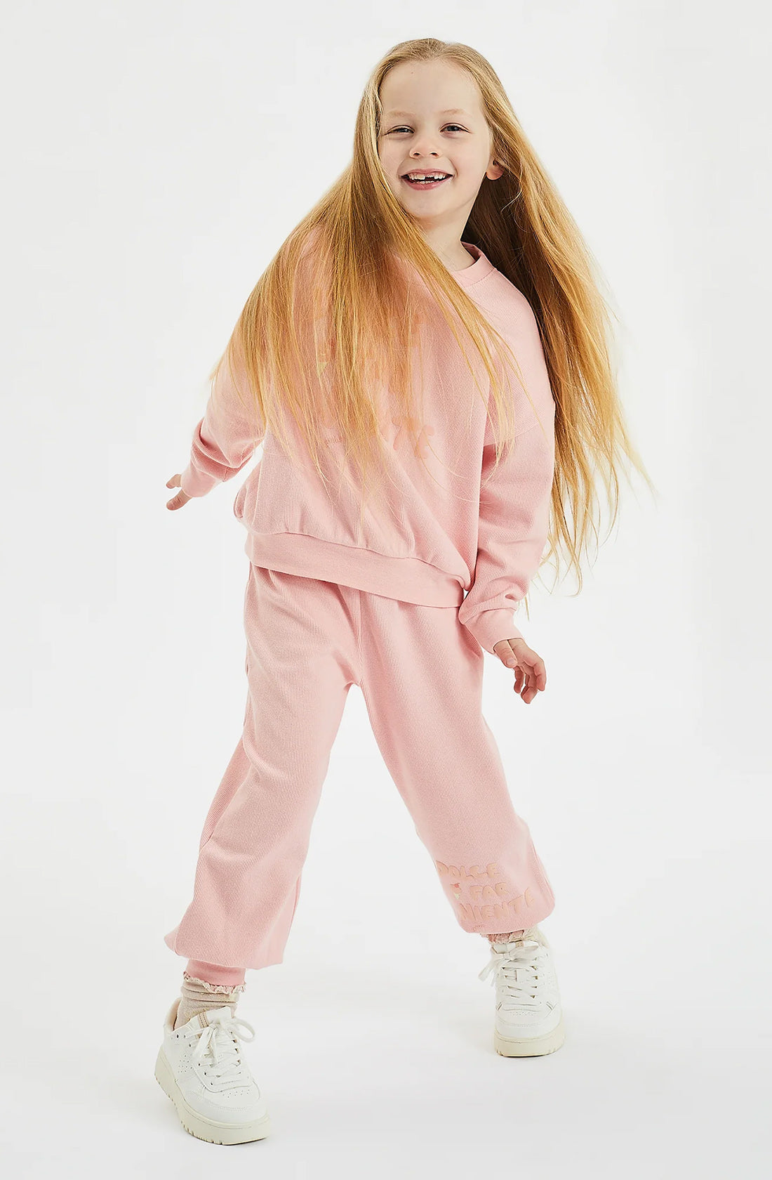 A child with long blonde hair smiles and poses playfully, wearing a long-sleeved pink sweatshirt and matching Dolce Lounge Pant Pink Lemonade by GOLDEN CHILDREN in a relaxed fit. They are also wearing white sneakers. The background is plain white.