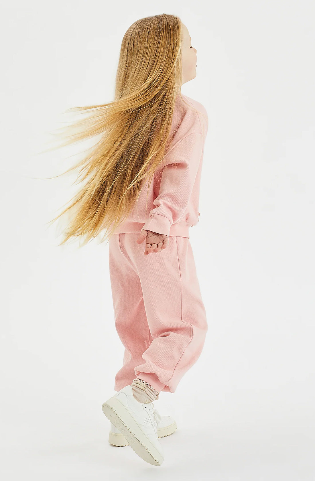 A young girl with long, flowing blonde hair is captured mid-jump against a plain white background. She is wearing a matching light pink sweatshirt and relax-fit Dolce Lounge Pant Pink Lemonade set from GOLDEN CHILDREN, along with white sneakers. The girl is facing sideways, with her hair and clothes appearing in motion.