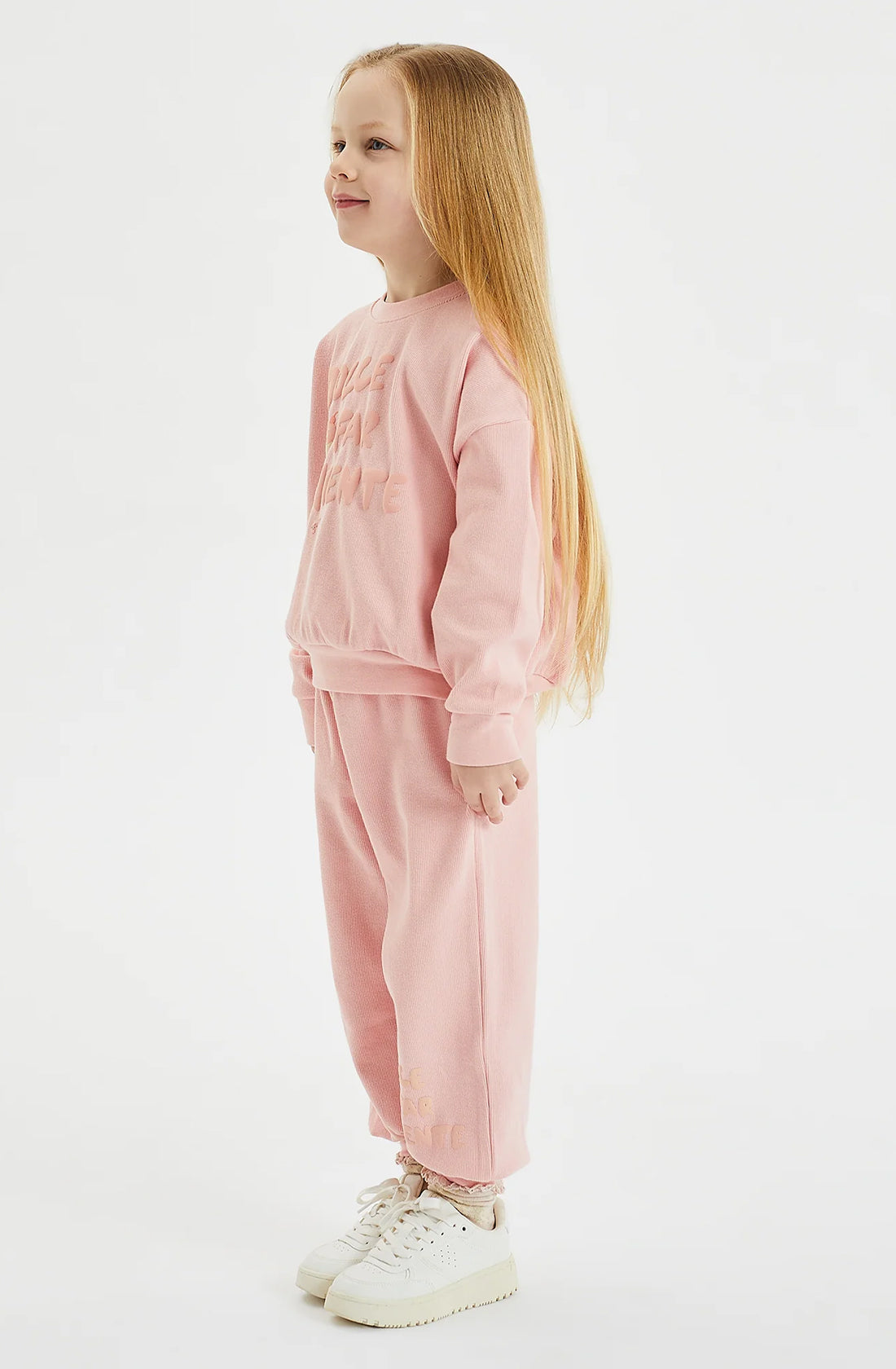 A young child with long blonde hair stands smiling, wearing a matching pink sweatshirt and GOLDEN CHILDREN's Dolce Lounge Pant Pink Lemonade set made from 100% cotton rib, paired with white sneakers. The relaxed fit outfit complements the child's joyful expression as they stand against a plain white background, giving a profile view to the camera.