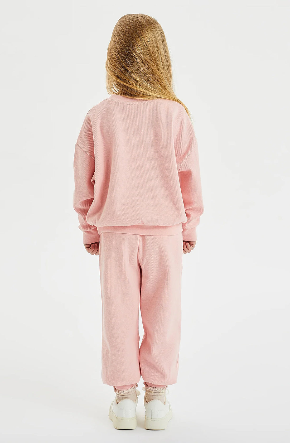 A young child with long blonde hair stands facing away from the camera, dressed in a matching light pink sweatshirt and GOLDEN CHILDREN's Dolce Lounge Pant Pink Lemonade in a relaxed fit. Their white sneakers complete the look against the plain white background. The outfit, made from 100% cotton rib, offers comfort and style.