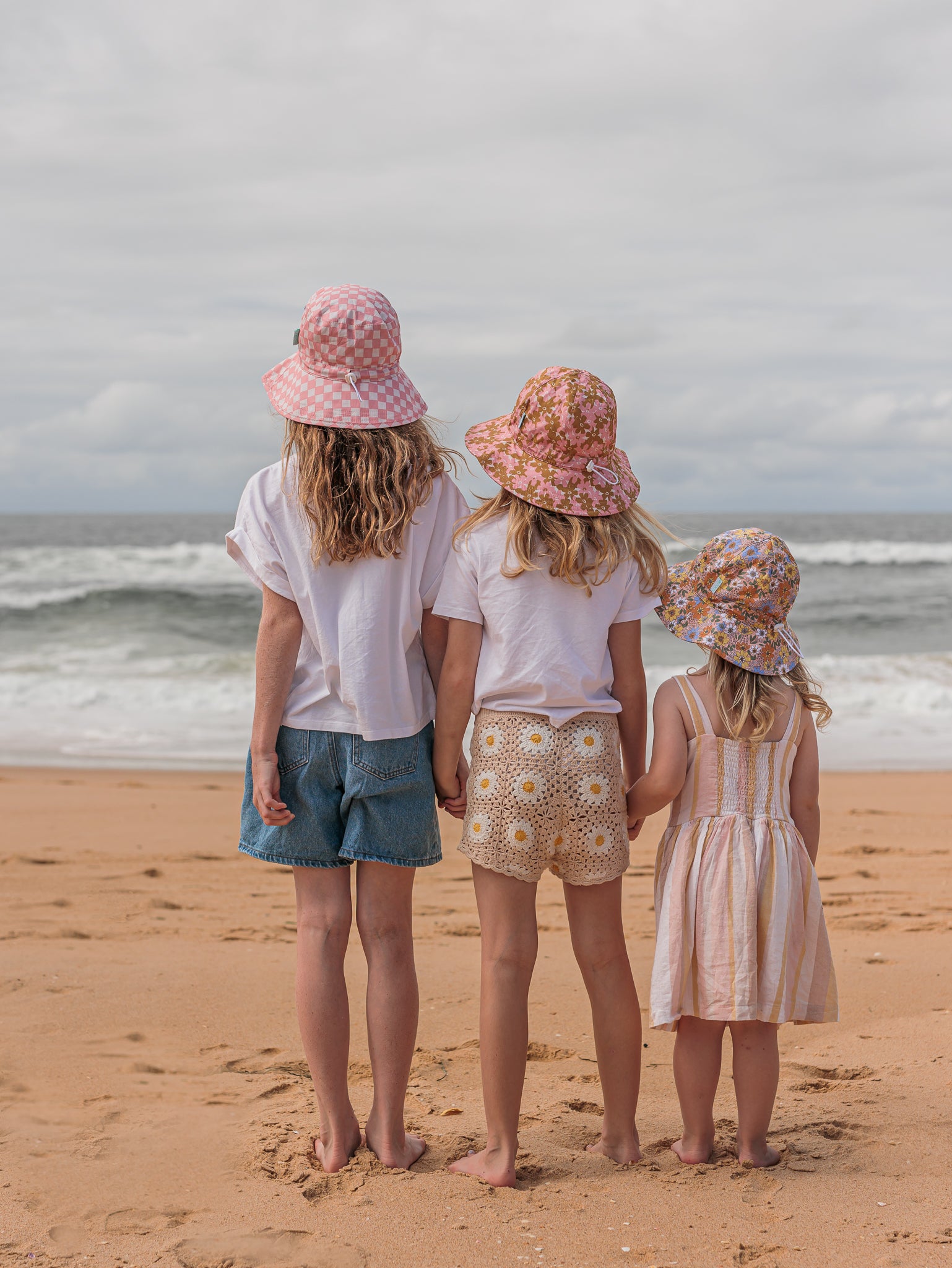 Three children stand on a sandy beach, facing the ocean. They wear cotton clothing and Pink Checks Broad Brim Bucket Hats by ACORN KIDS, which offer UPF50+ sun protection. The sky is cloudy, and the waves gently crash on the shore.