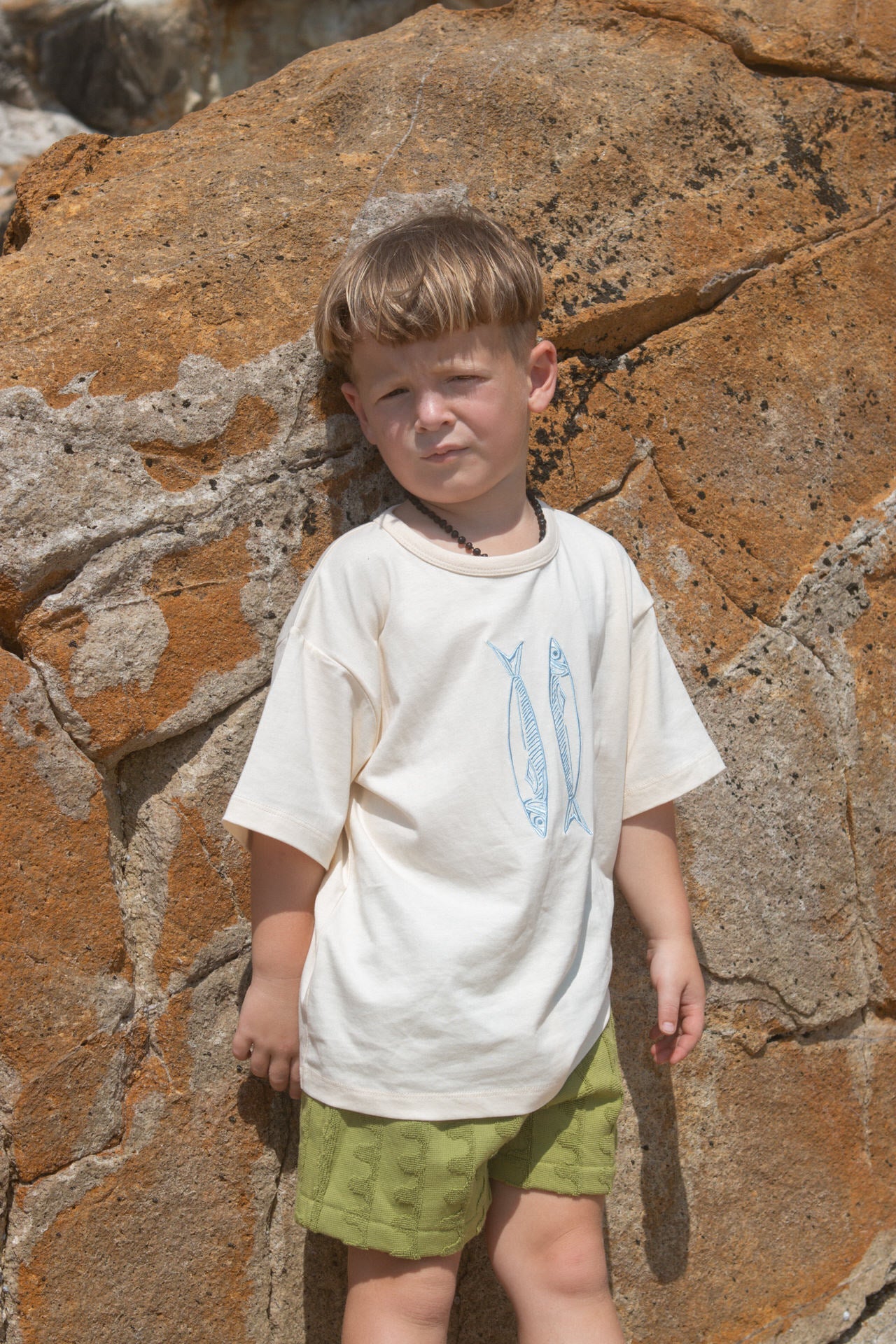 Child wearing a white t-shirt with blue fish and green shorts standing against a rocky wall.