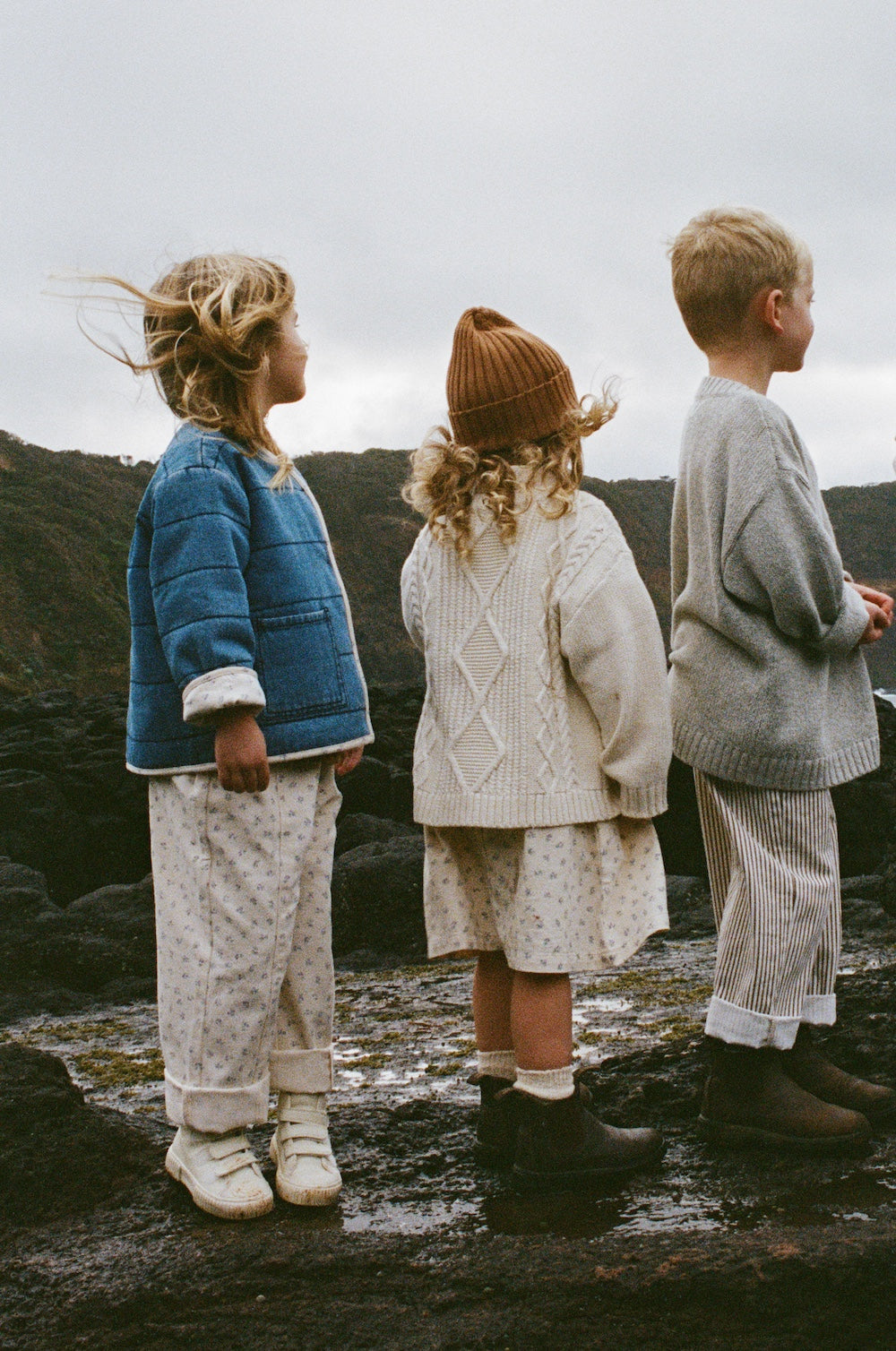 Three young children stand outdoors on rocky ground, dressed in cozy GROWN Collar Dress Garden outfits. The sky is overcast, with hills visible in the background.