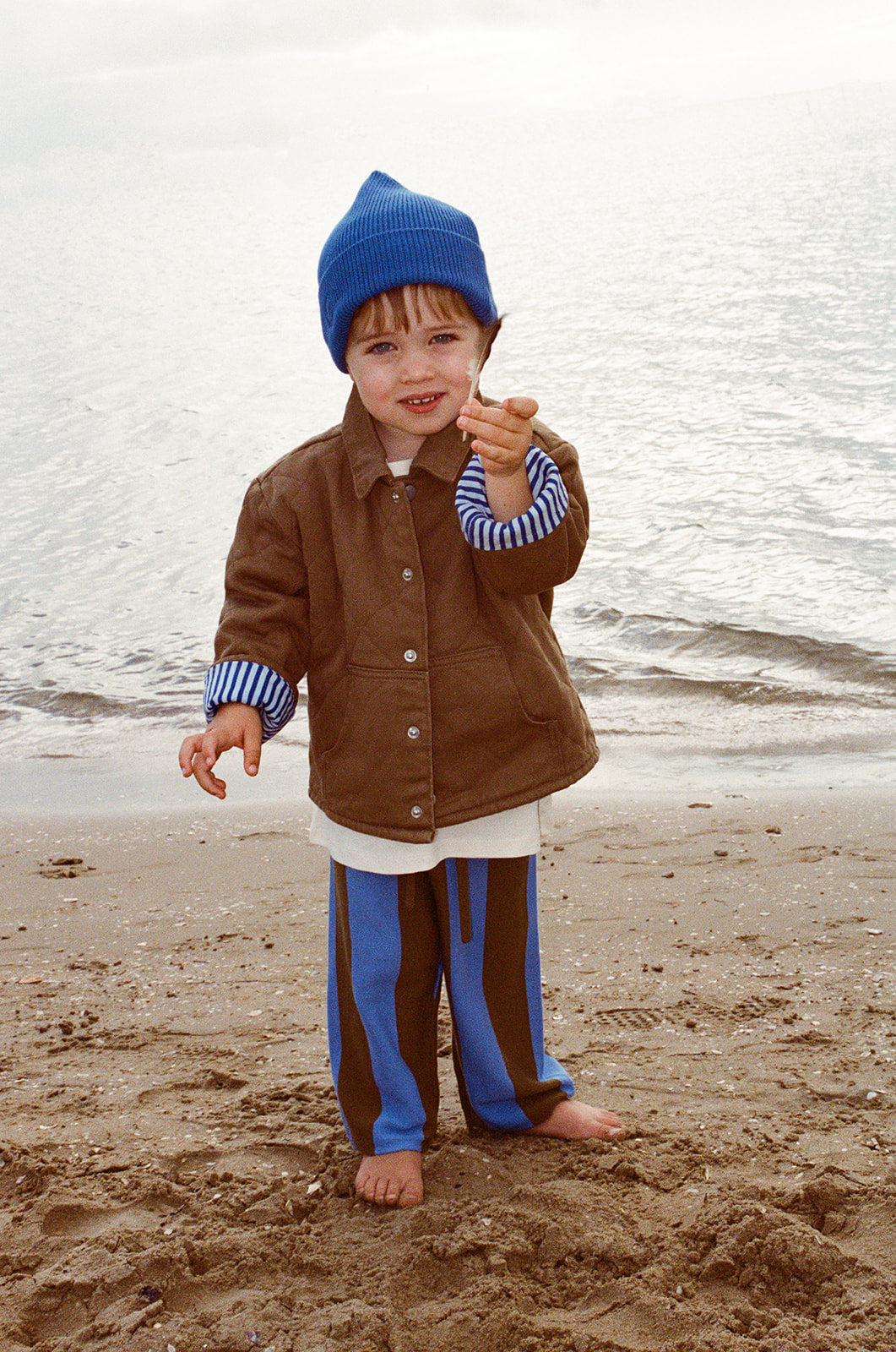 A young child wears the GROWN Organic Quilted Denim Jacket Coffee, standing barefoot on a sandy beach in front of the ocean, smiling and pointing towards the camera.