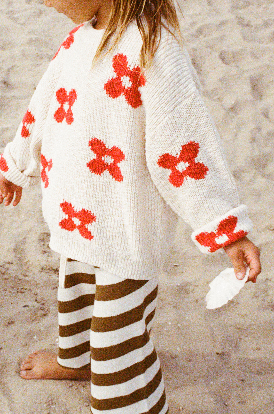 A child stands on sand wearing GROWN’s Diagonal Stripe Knit Flares—brown-and-white striped pants with an elastic waistband—and a white sweater with red flower patterns, holding a white seashell.