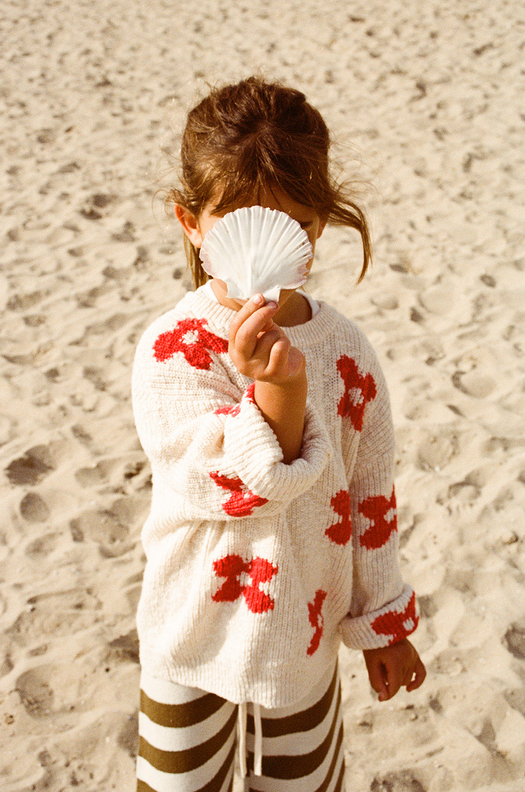 A child wearing the GROWN Knitted Posy Pull Over stands on a sandy beach, holding a white seashell in front of their face.