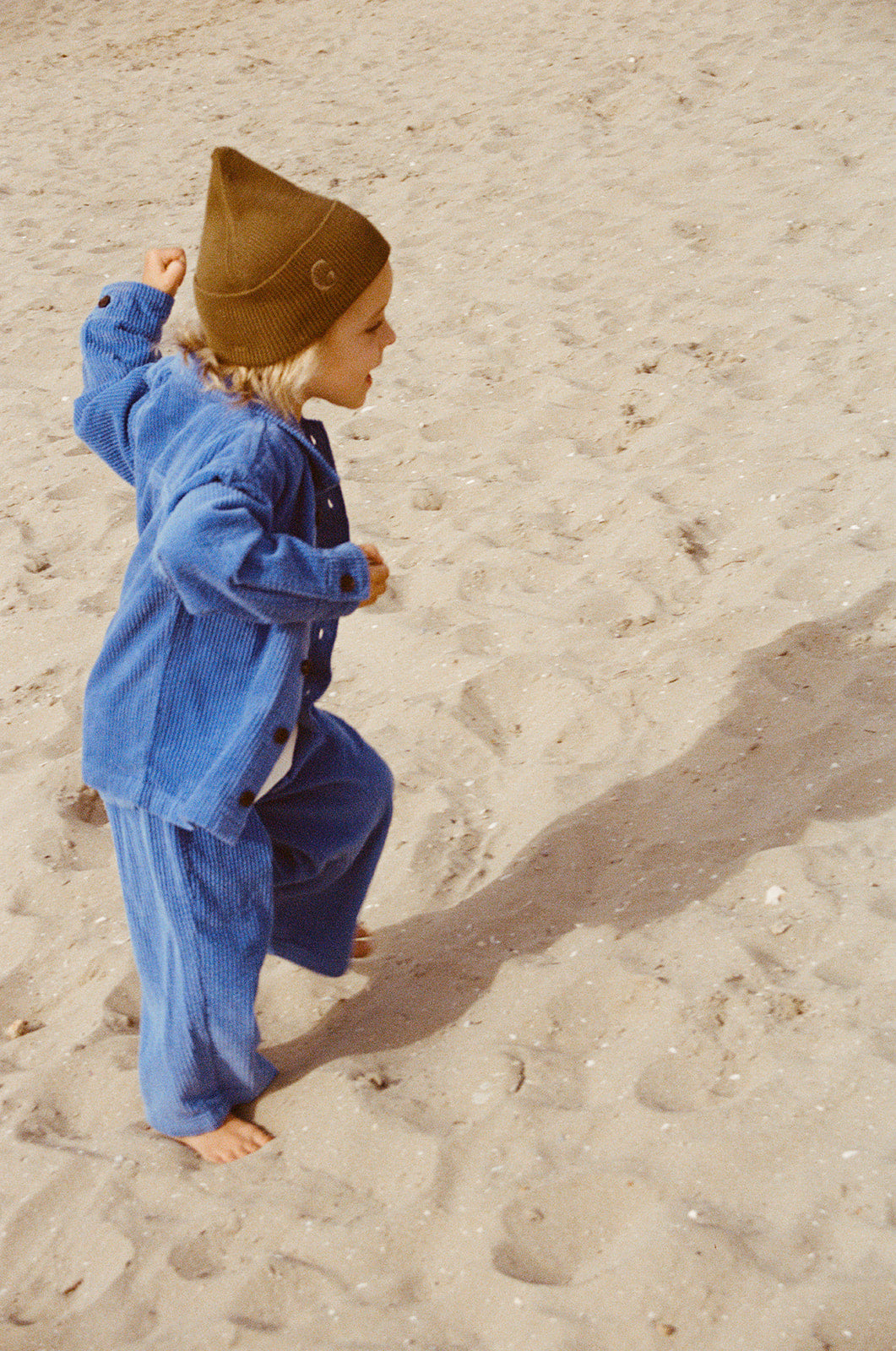 A young child in GROWN's Organic Relaxed Cord Pant Sea joyfully runs barefoot on sandy ground, wearing a brown beanie and casting a sunlit shadow.