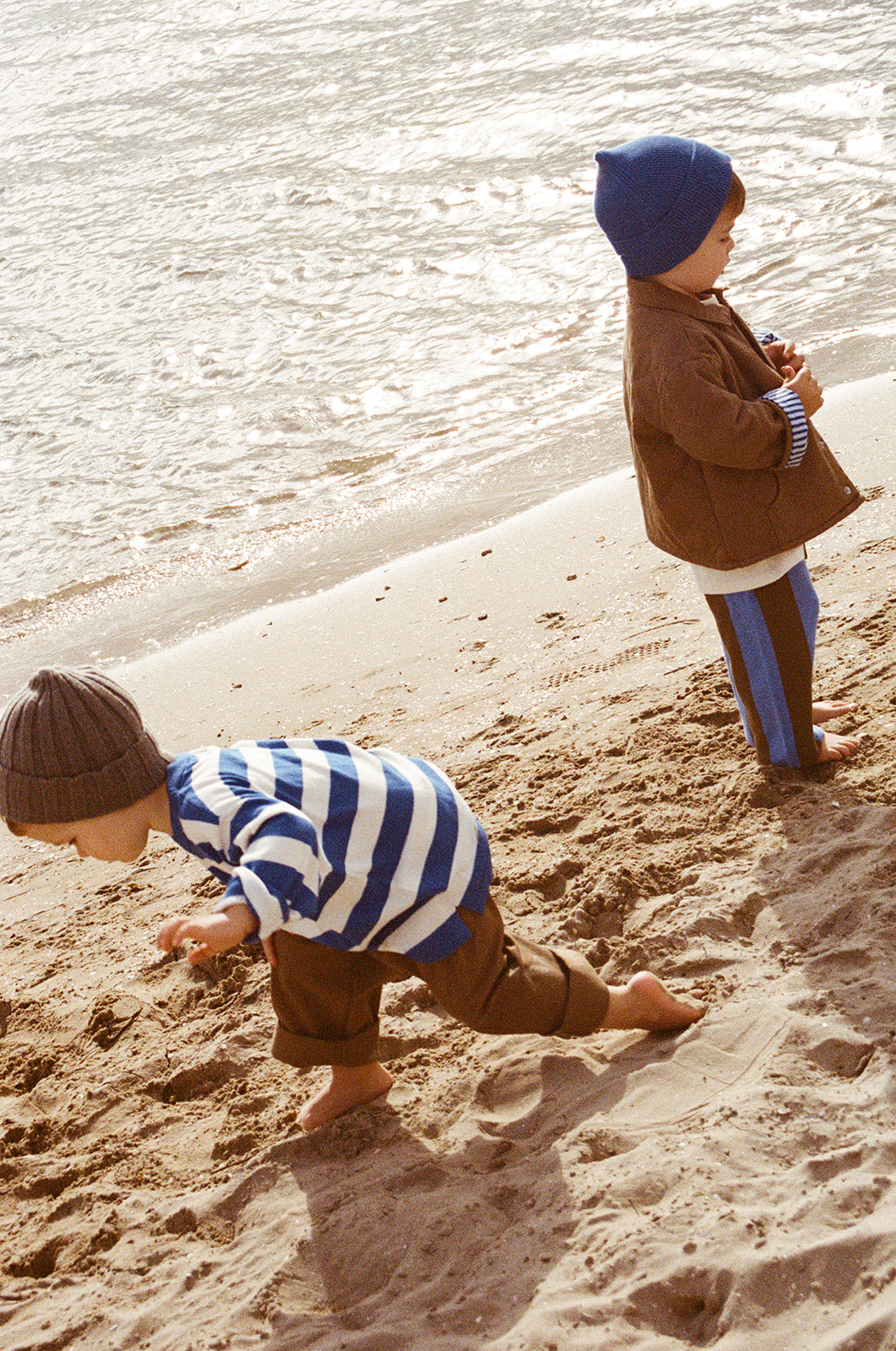 Two young children play barefoot on a sandy beach near the water; one bends down to touch the sand while the other stands, both wearing GROWN - Stripe Long Sleeve Tee by GROWN.