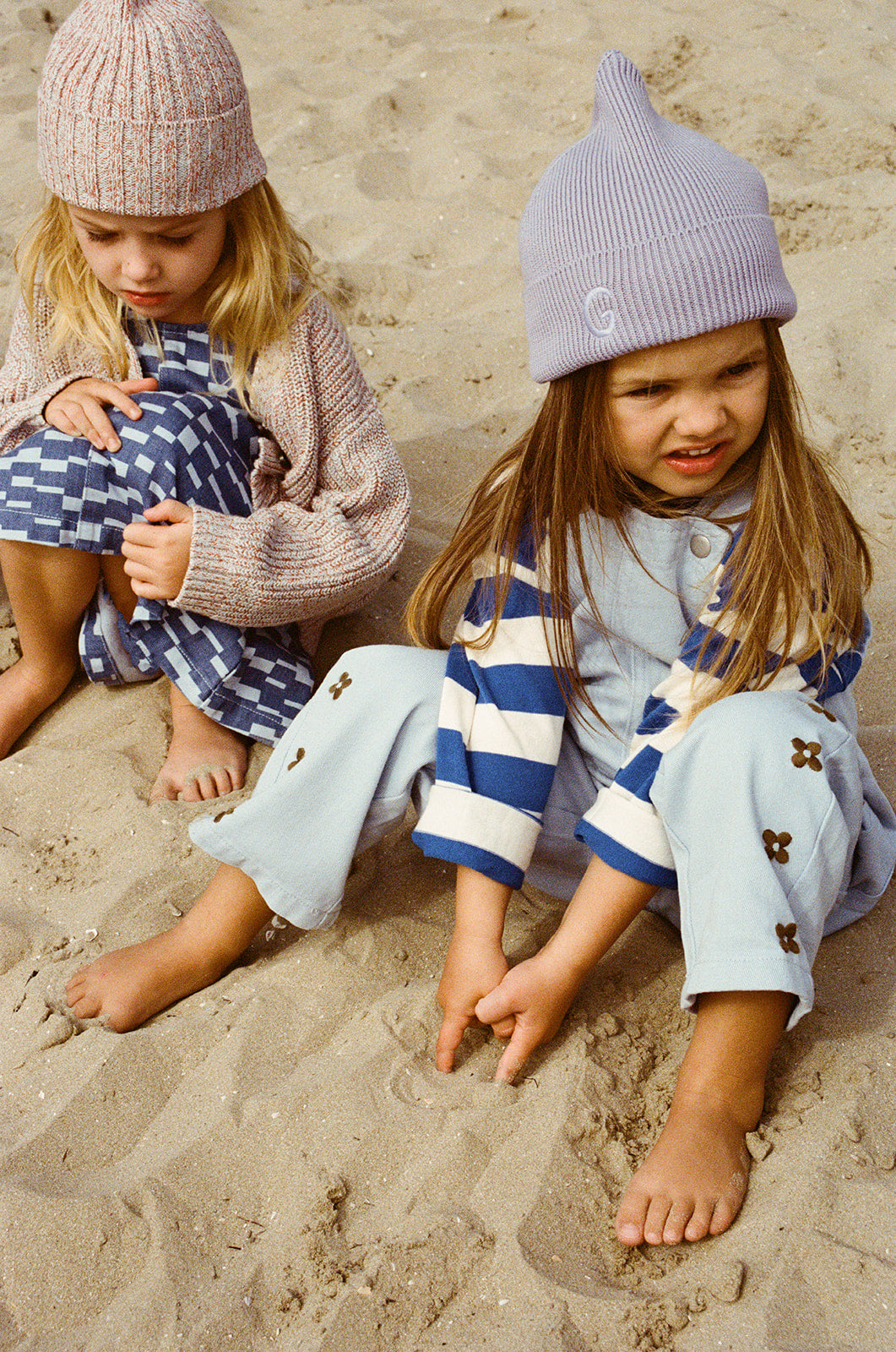 Two young children sit barefoot on the sand. One smiles, wearing GROWN’s Organic Embroidery Posy Overalls, while the other looks down in a blue patterned dress and knit hat.