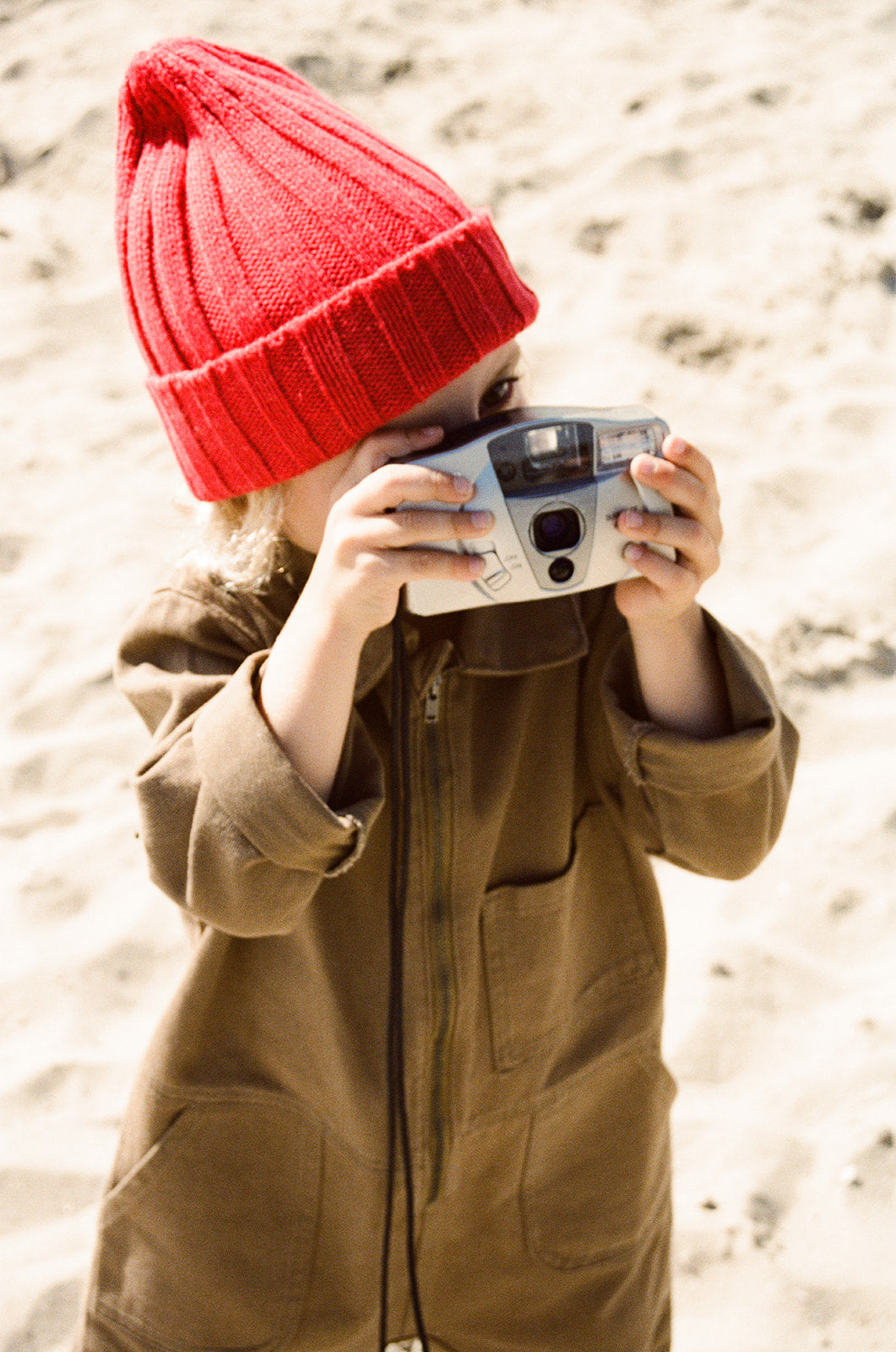 A child in the GROWN Merino Blend Ribbed Beanie Flame, a cozy red hat, lifts a camera to their face while standing on a sandy beach.