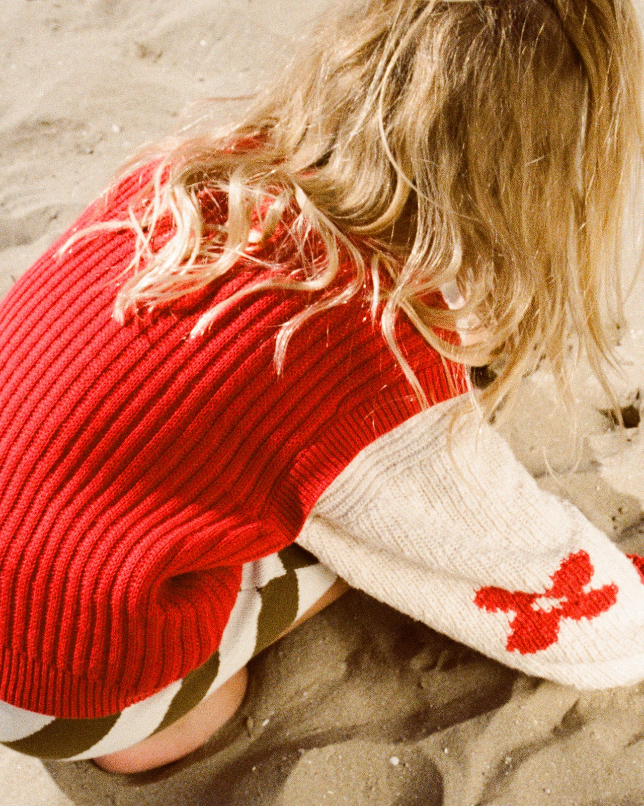 A child with long blonde hair kneels on sand, wearing the GROWN Chunky Rib V-Neck Vest Flame. Made from organic cotton for comfort, the oversized-fit vest features a red design on the sleeve. The child's face is not shown.