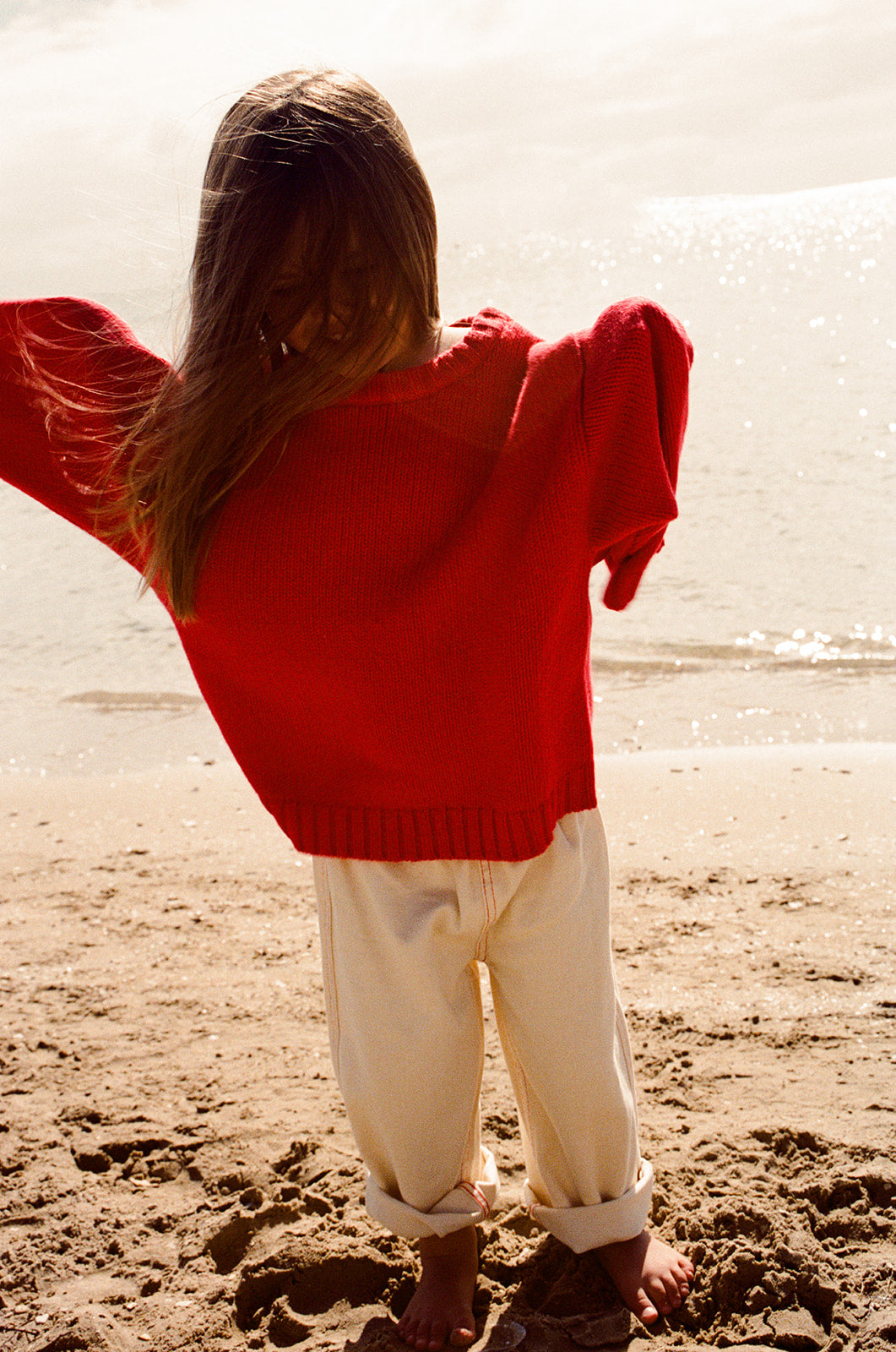 A child with long hair wearing the GROWN Merino Blend Everyday Pull Over Flame stands barefoot on a sandy beach, ocean in the background, paired with rolled-up cream pants.