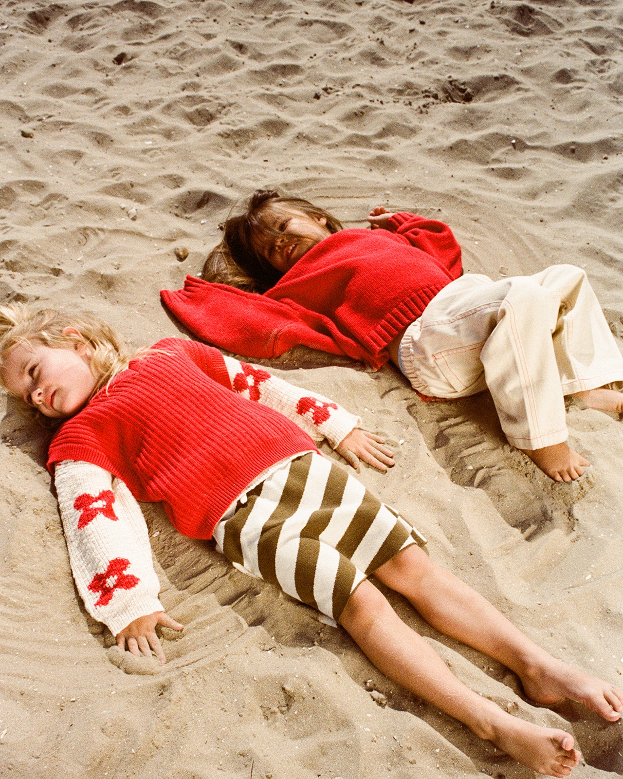 Two young children relax on the sand in cozy outfits. One styles a floral cardigan with striped shorts, while the other wears GROWN’s Organic Everyday Denim Pant in Milk, looking serene and enjoying a sunny beach day.