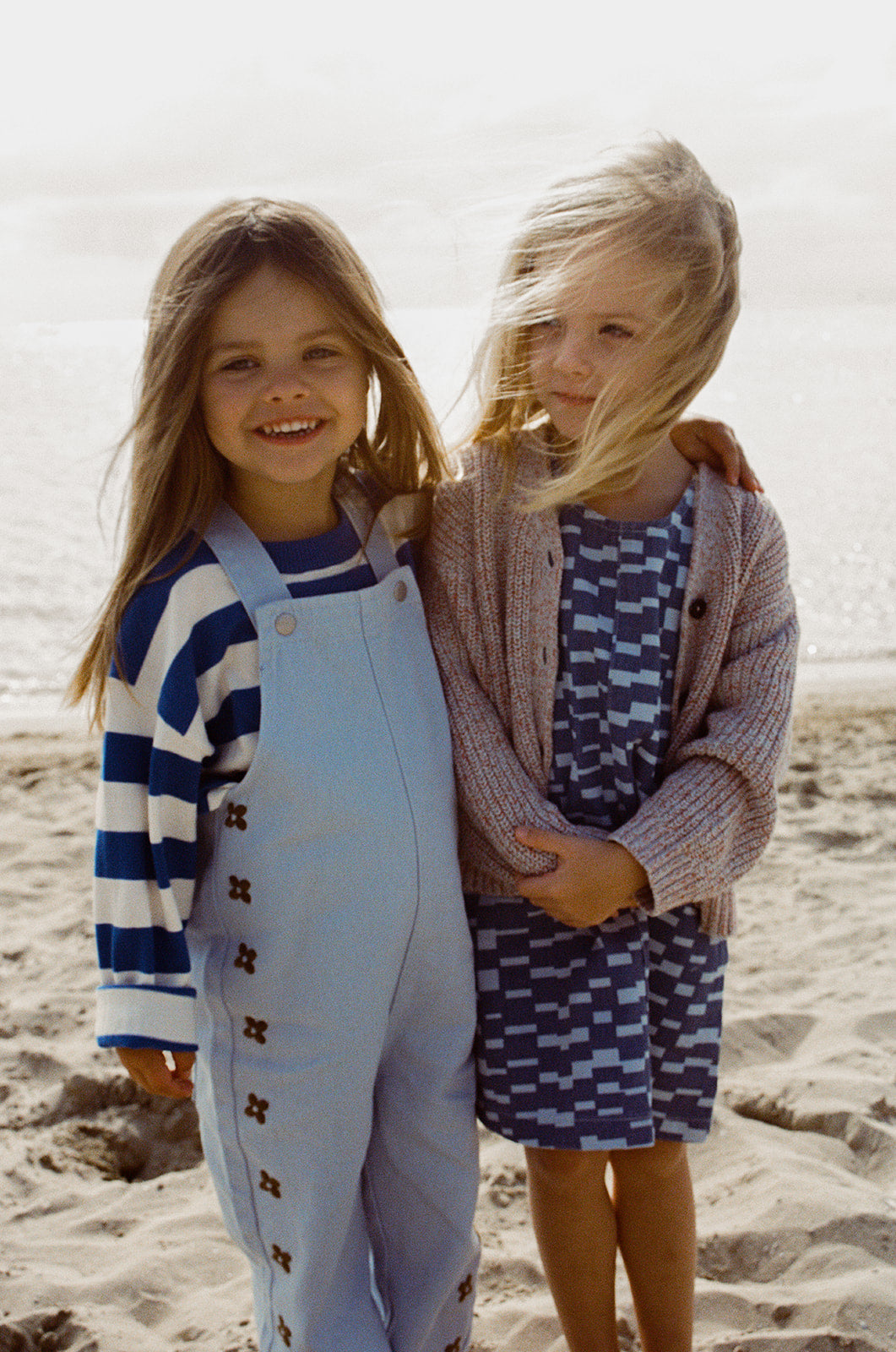 Two young children stand on a sandy beach. One smiles at the camera in light blue overalls and a striped shirt. The other wears the GROWN Boxes Denim Dress by GROWN with an A-line shape and cardigan, wind blowing her blond hair across her face.