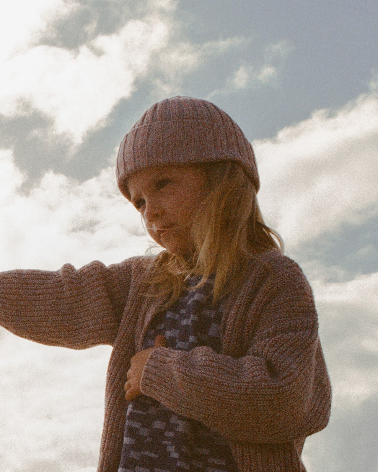 A young child with long hair, wearing the GROWN Twisted Chunky Rib Cardigan and a knit hat, stands outdoors against a cloudy sky, gazing to the side with a gentle expression.