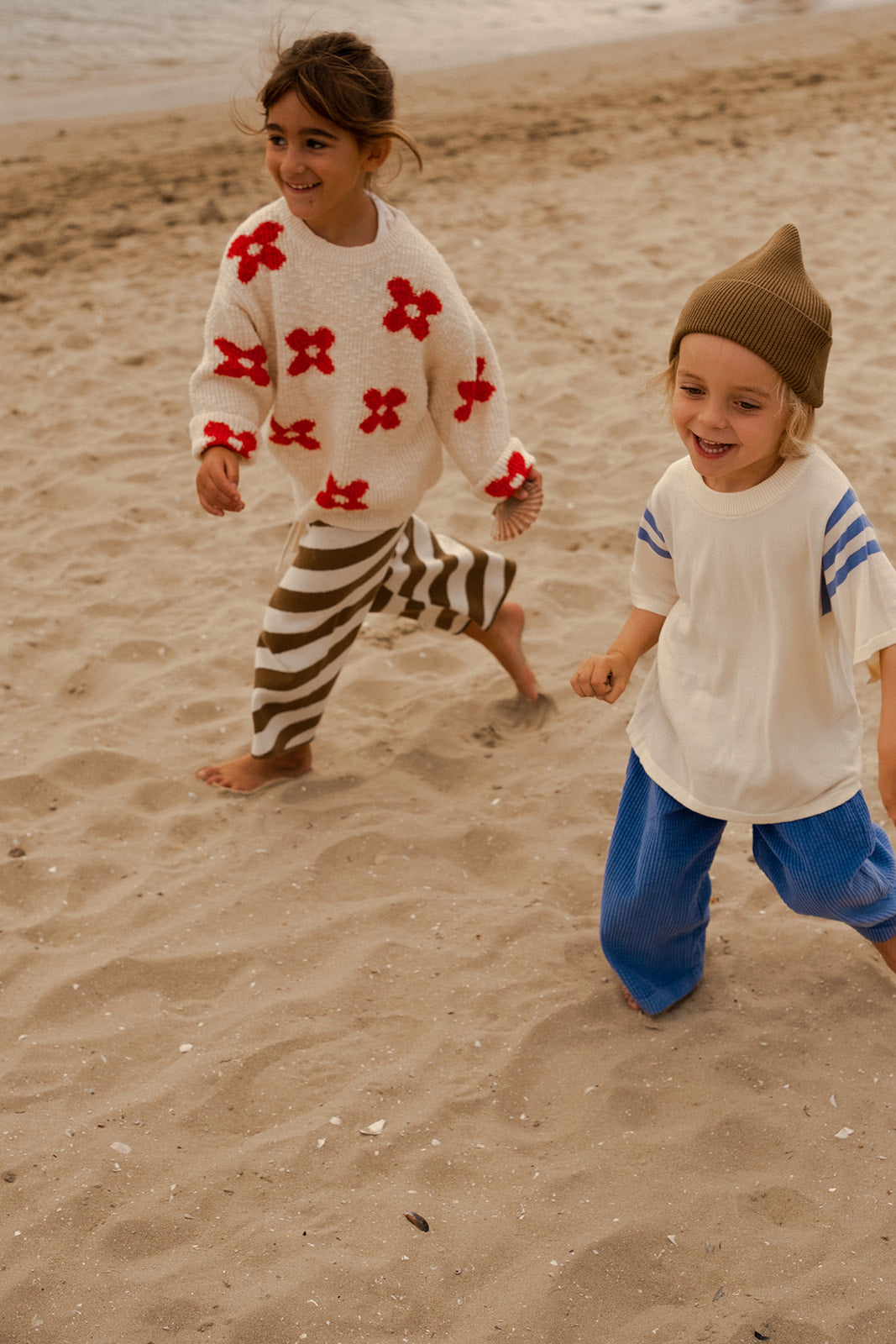 Two young children run barefoot on a sandy beach. One wears GROWN's Organic Relaxed Cord Pant Sea, while the other is dressed in a white sweater with red flowers and striped pants. The sea glistens in the background.