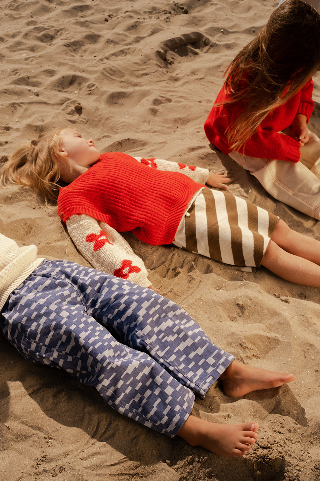 Three kids relax on sandy ground in cozy outfits. One wears the GROWN Chunky Rib V-Neck Vest Flame, another sports blue patterned pants with a cream organic cotton sweater, and the third mixes red and tan pieces.