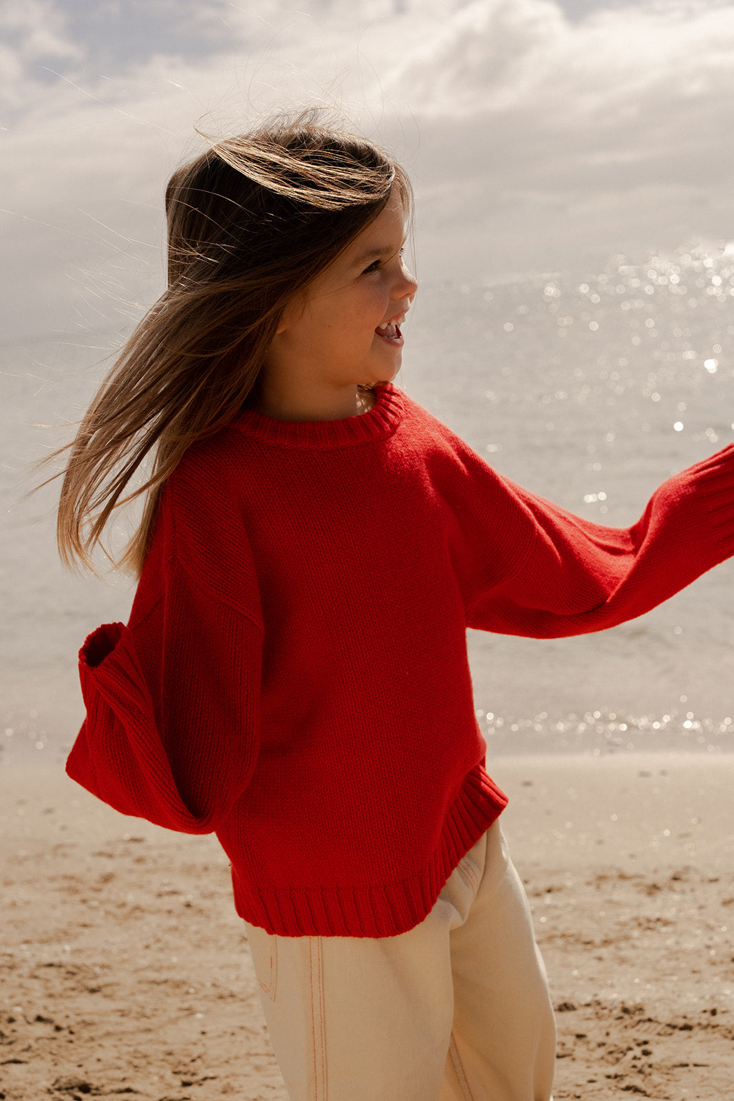 A young girl with long hair smiles on a sunny beach, wearing GROWN's Merino Blend Everyday Pull Over in Flame and light pants.